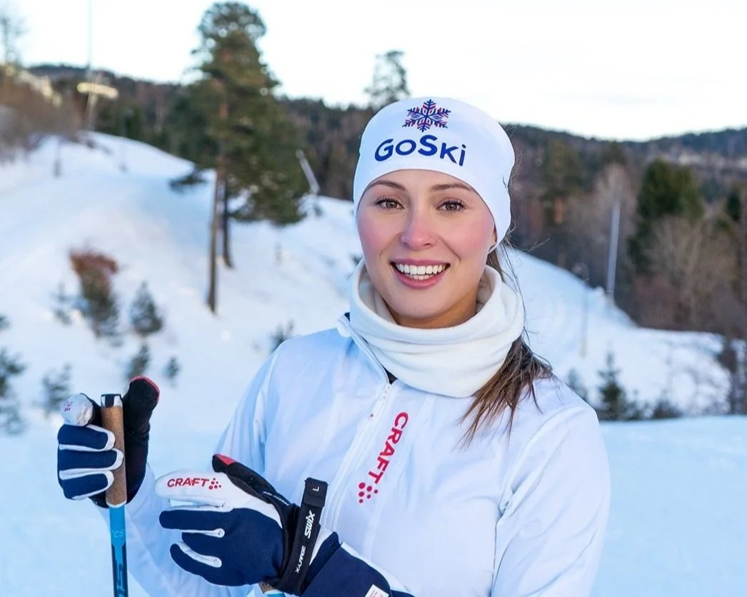Smilende kvinne i hvit jakke med blå og røde striper på ermene, svarte hansker og hvite håndflater, stående på en snødekt bakke med ski og staver. Personen har på seg en hvit lue med design detaljer. Bakgrunnen viser en klar blå himmel og trær.
