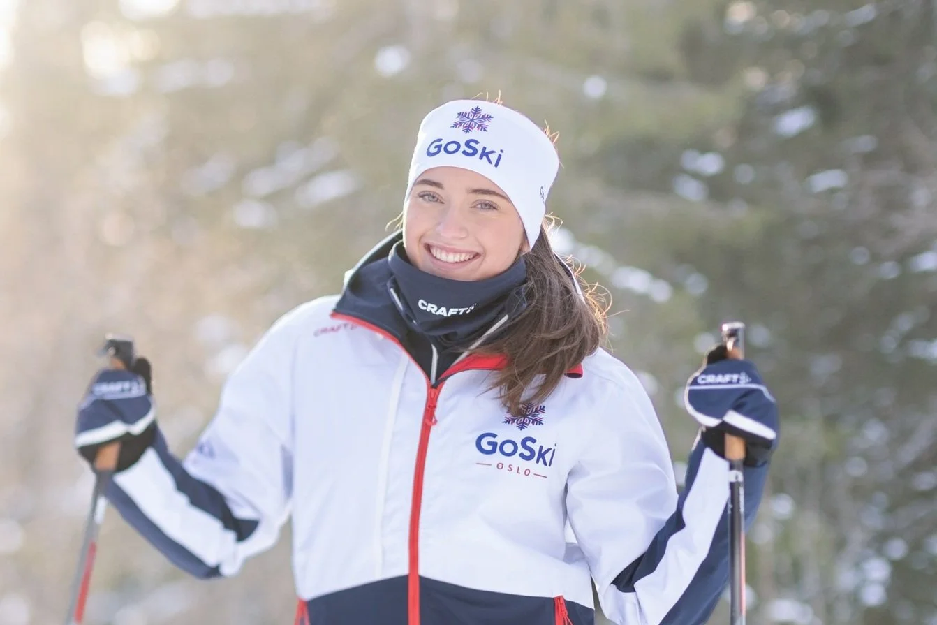 Skiinstruktør i profesjonell antrekk med skistaver, stående på et snødekt landskap med klar himmel og furutrær i bakgrunnen, med logoen ‘GoSki’ synlig på jakken.