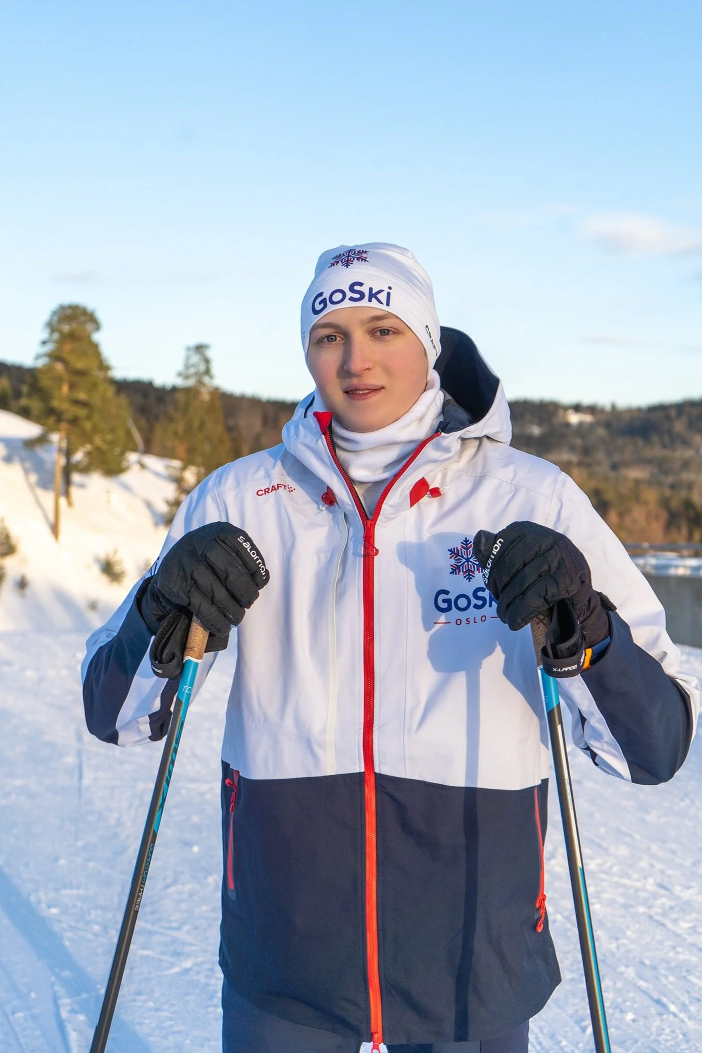 Skiinstruktør i profesjonell antrekk med skistaver, stående på et snødekt landskap med klar himmel og furutrær i bakgrunnen, med logoen ‘GoSki’ synlig på jakken.