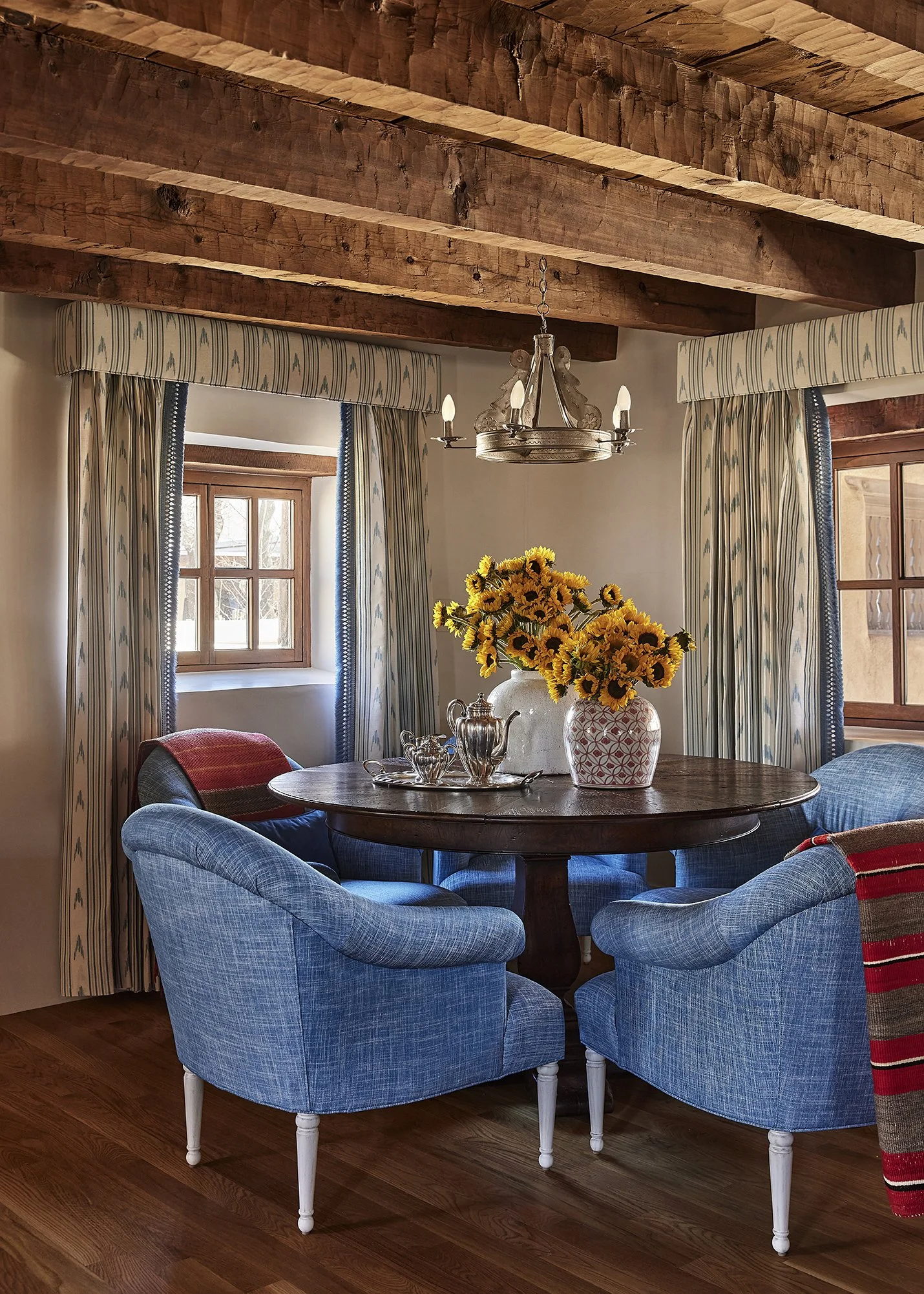 Round wooden dining table surrounded by blue upholstered chairs, with a large vase of sunflowers as the centerpiece beneath rustic wood ceiling beams.