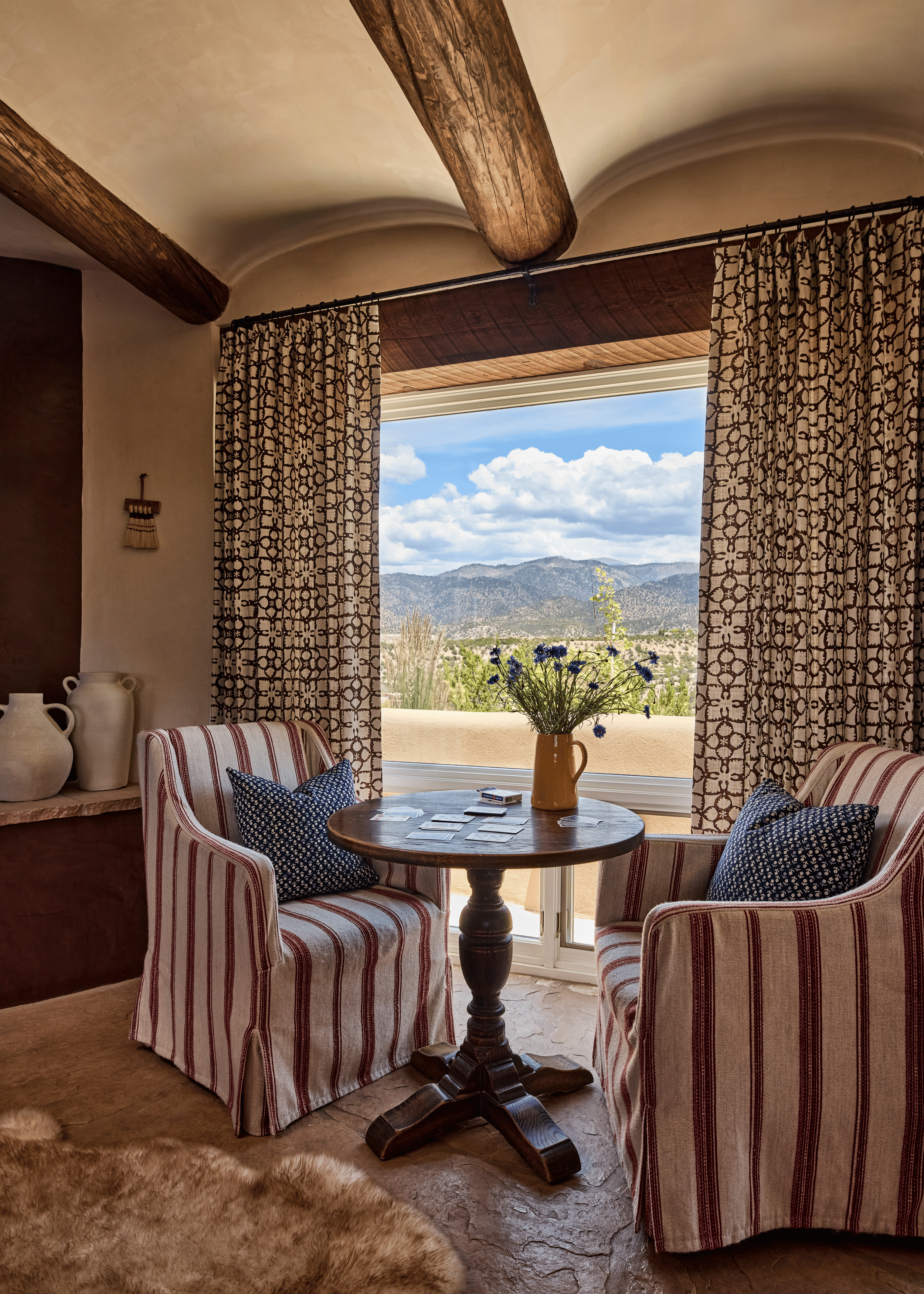 A sitting nook designed around the view: two red-stripe slipcovered chairs, a round pedestal table, and block-print curtains that give just enough frame to the Sangre de Cristo Mountains stretching beyond. French & French Interiors placed this corner