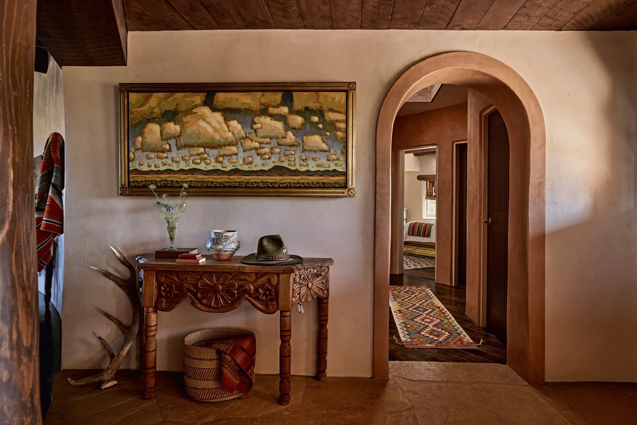 Looking back through the entry hall toward the living room, the Mesa Project shows its full depth: a carved console with Pueblo pottery, elk antlers propped against the wall, and the green velvet sofa visible beyond. French & French Interiors treats 