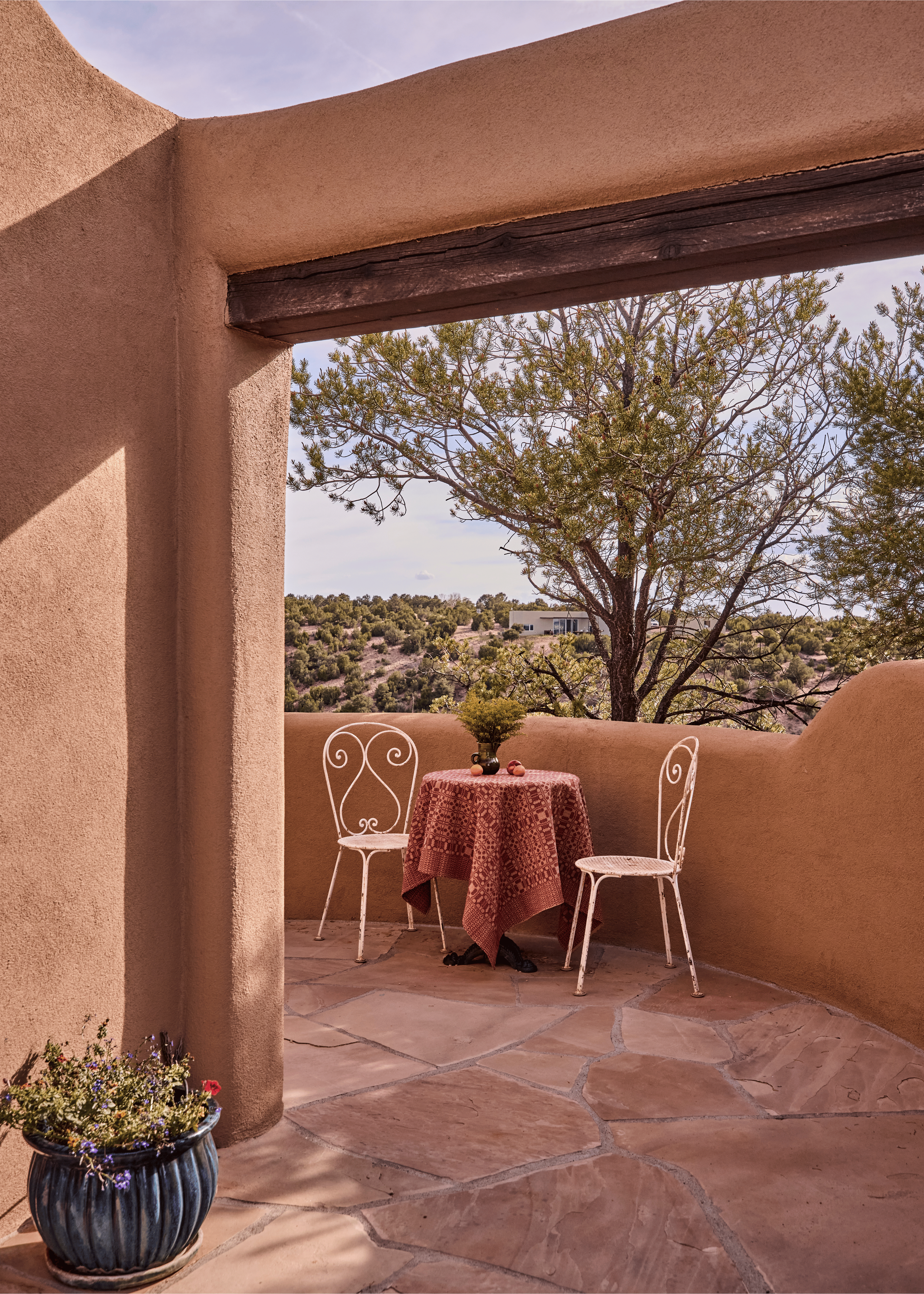 A covered portal at the Mesa Project frames the high desert landscape of Tesuque, New Mexico - adobe architecture at its most elemental, styled by French & French Interiors with a patterned cloth and vintage ironwork chairs that feel entirely at home