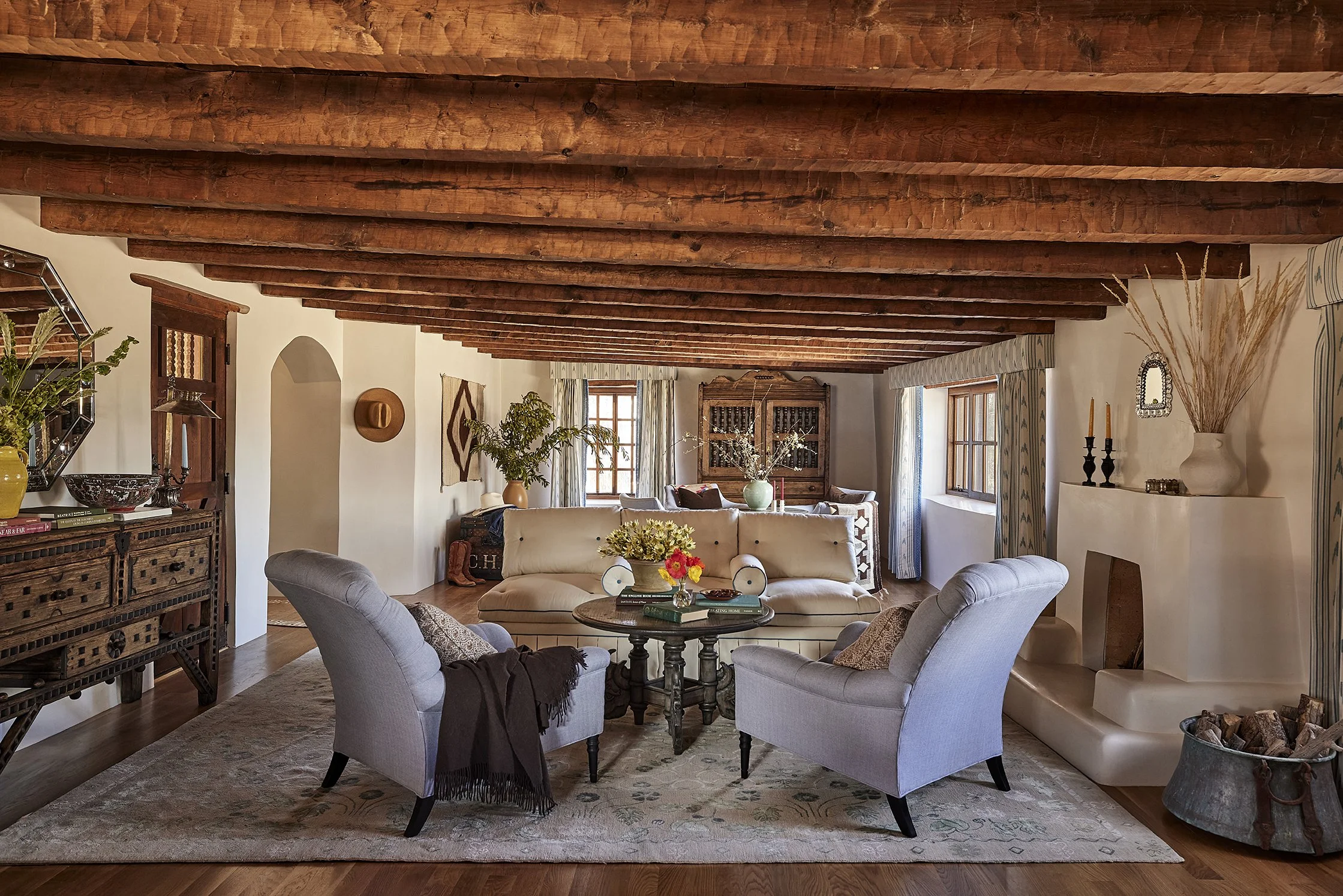 A spacious living room with wood-beamed ceilings, cream sofas, and pale lavender armchairs arranged around a central round table. A white plaster fireplace, rustic console, and large rug add to the Southwestern charm.