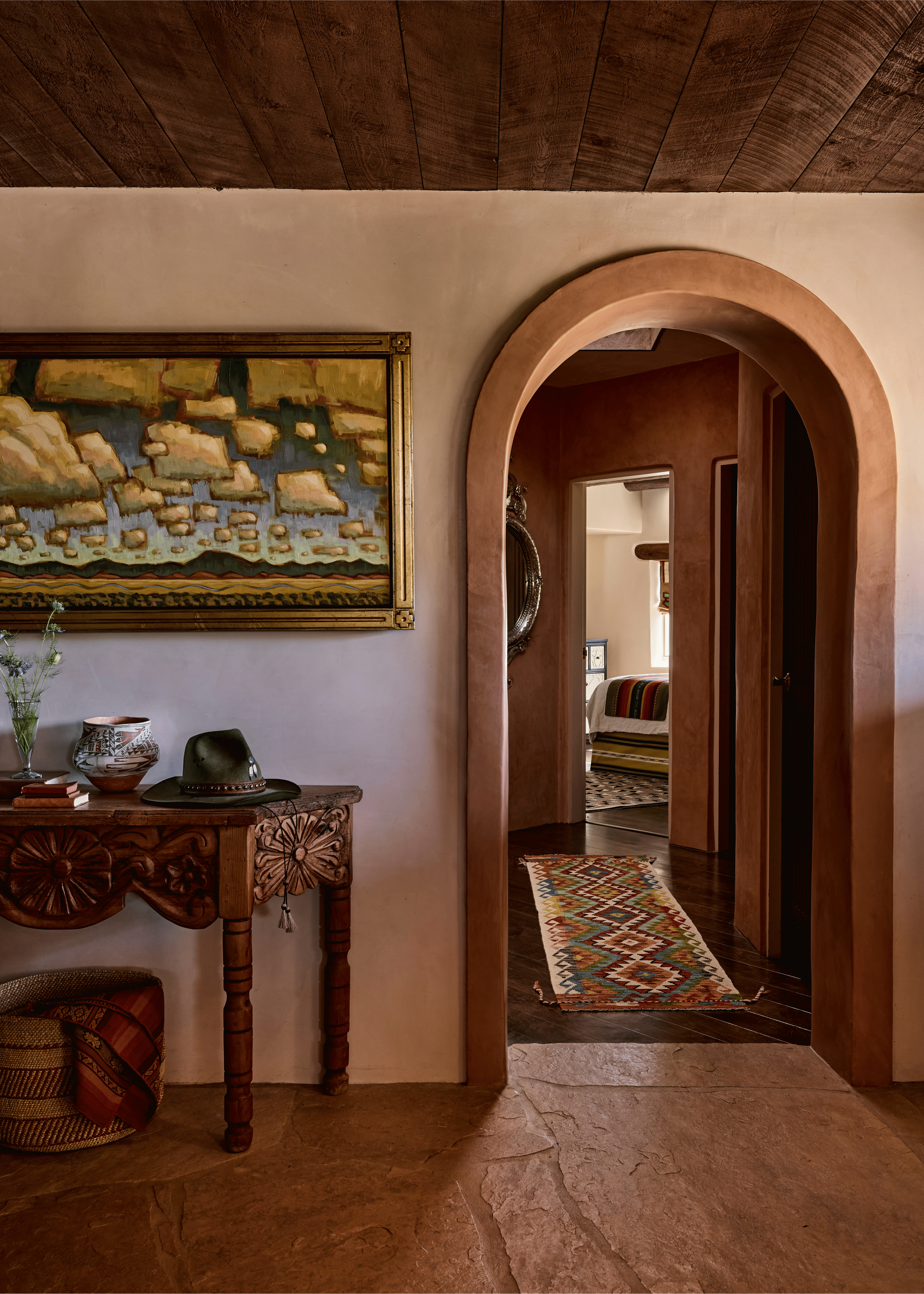 A curved adobe banquette upholstered in patchwork fabric anchors one end of the Mesa Project's living room, where a bold figurative oil painting and deep red built-in cabinetry create a corner that feels as curated as a gallery and as comfortable as 