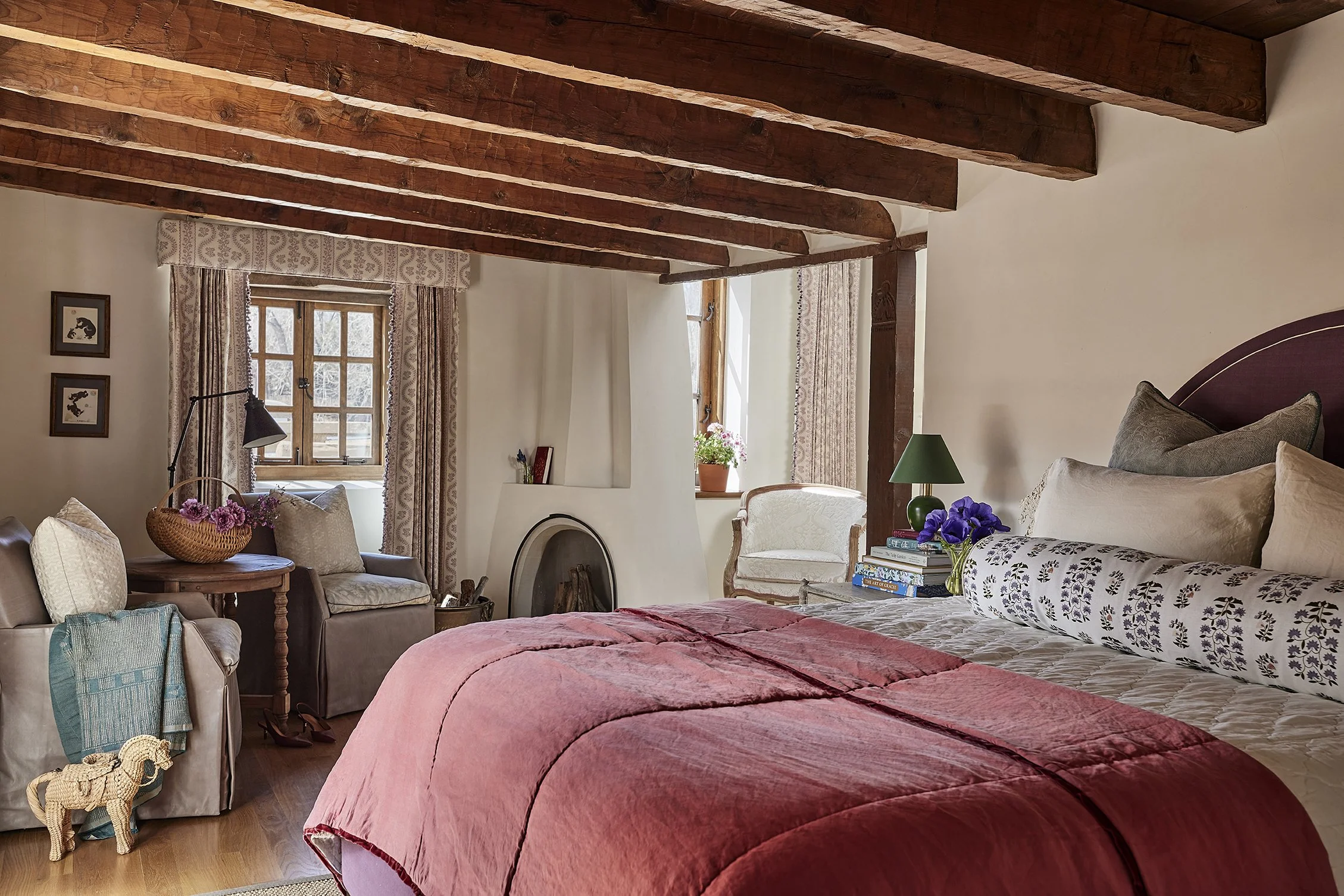 A cozy bedroom with exposed beams, a bed dressed in a bold red quilt and patterned bolster pillow, and seating by a plaster fireplace. Layered textiles and potted plants bring warmth to the space.