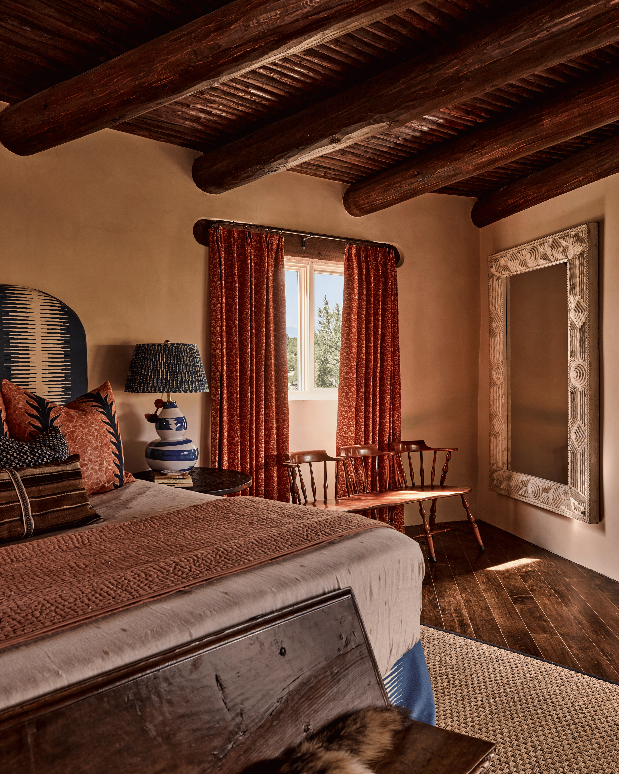The full bedroom at the Mesa Project reads like a carefully edited collection: a navy arched headboard, an antique carved mirror leaning against the wall, rust block-print drapes filtering desert light, and a writing desk tucked against the window. D