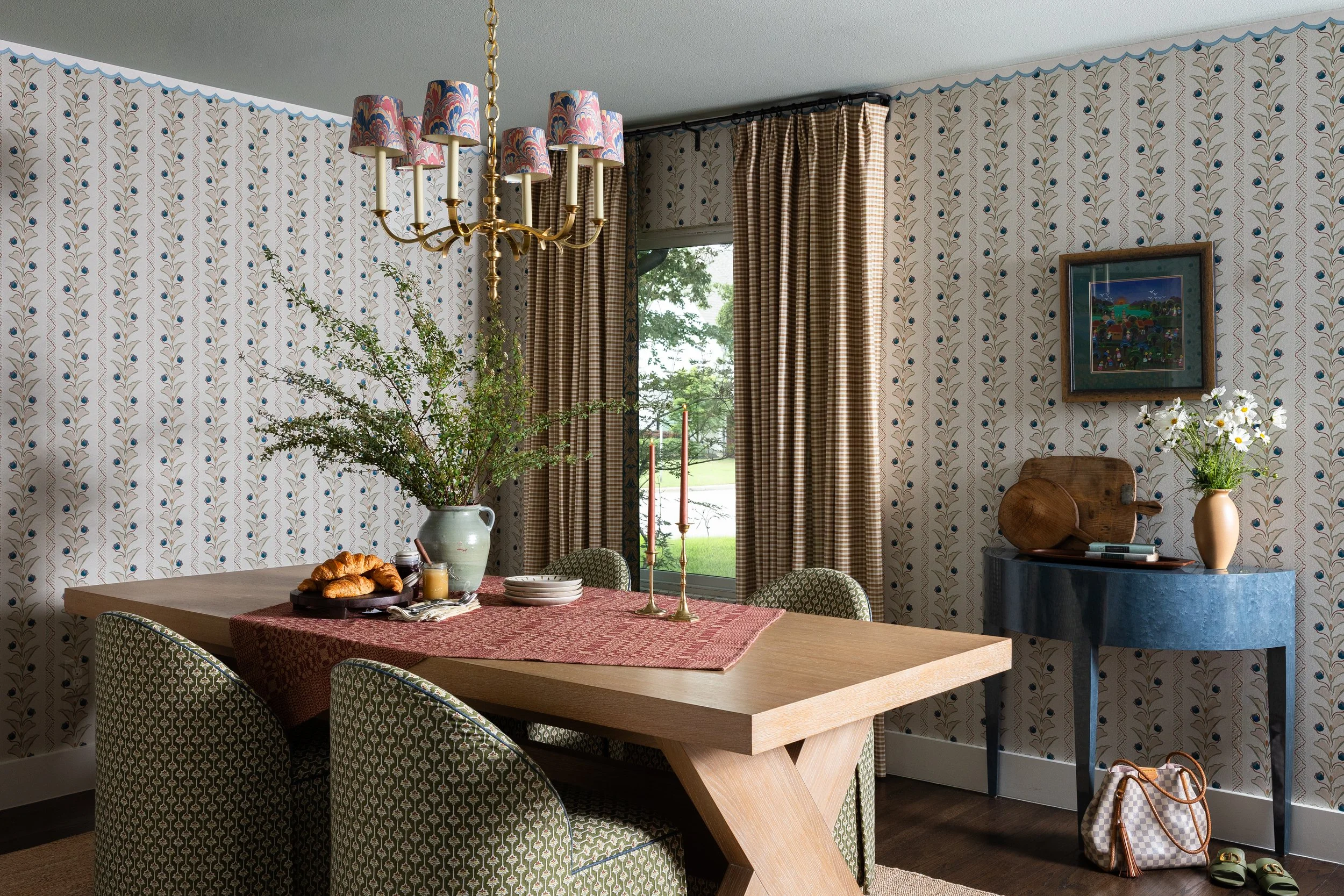 A dining room in a Dallas luxury cottage interior - botanical wallpaper trimmed with a hand-painted scallop border, a brass chandelier fitted with marbled shades, and green upholstered chairs that invite long meals and slow mornings.