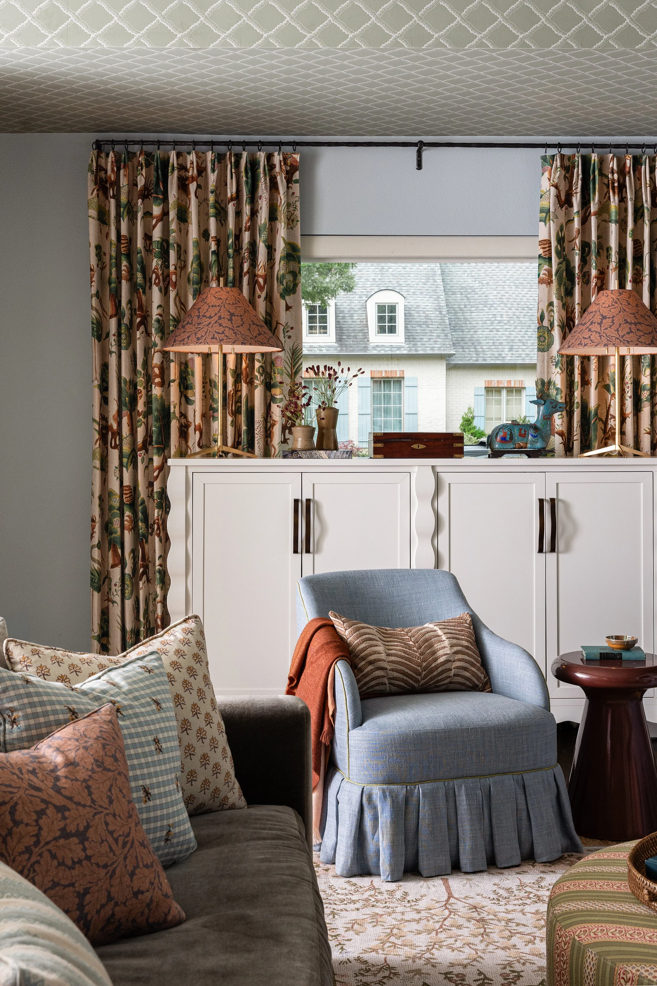 A skirted armchair layered with block print and gingham pillows sits beneath botanical drapery in this Dallas family home - every detail considered, nothing precious.