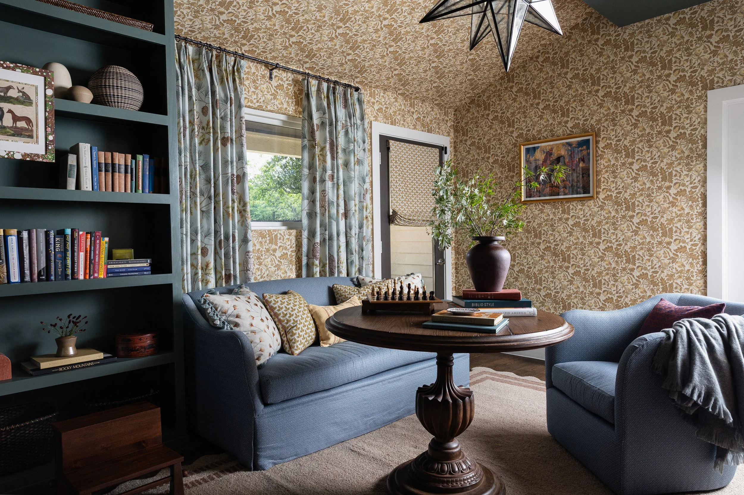 The sitting area of this Dallas cottage study - gold botanical wallpaper wrapping walls and ceiling, a carved pedestal table set with a chess board, and blue upholstered seating that invites long afternoons with books and good company.