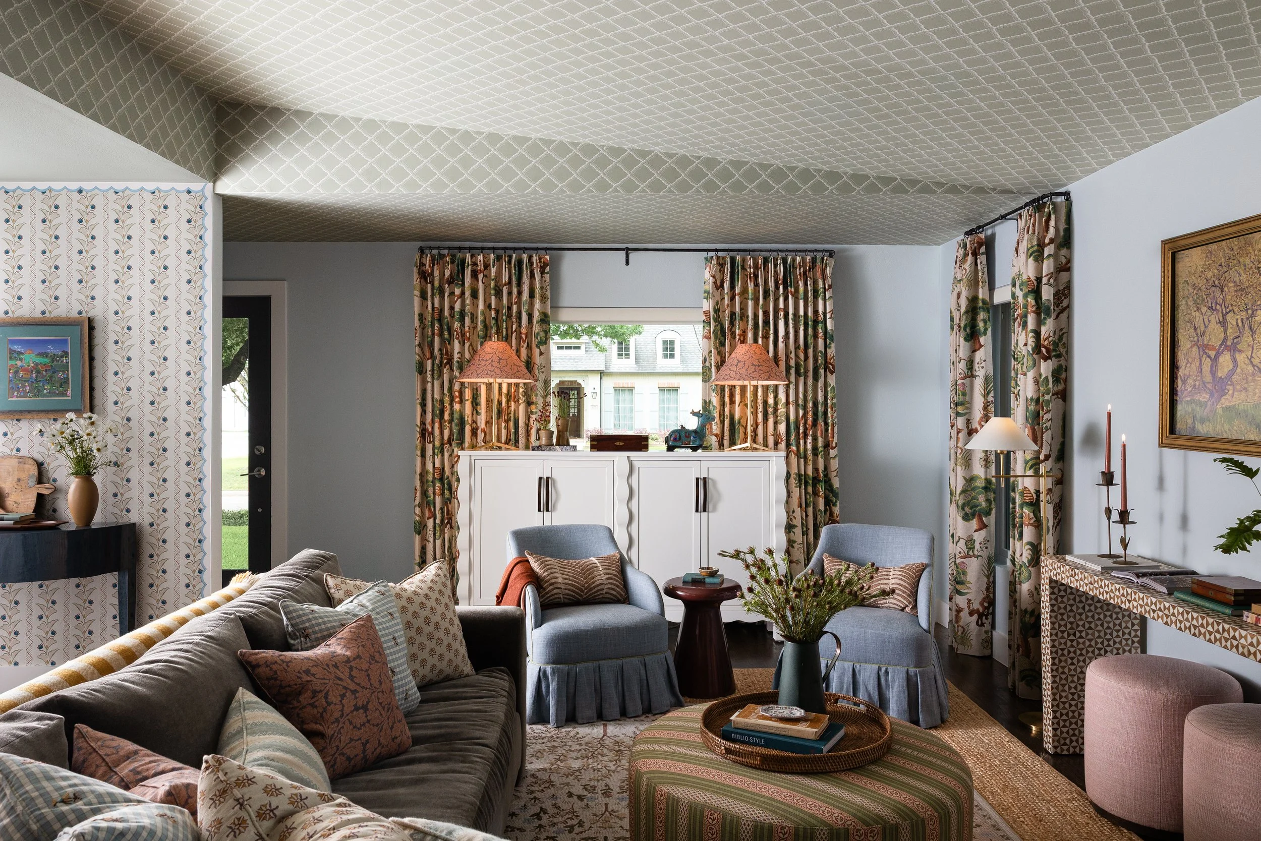 In this Dallas cottage interior, a wallpapered trellis ceiling anchors a living room layered with botanical drapery, mixed-print pillows, and skirted armchairs - a signature French and French approach to warm, collected luxury design.