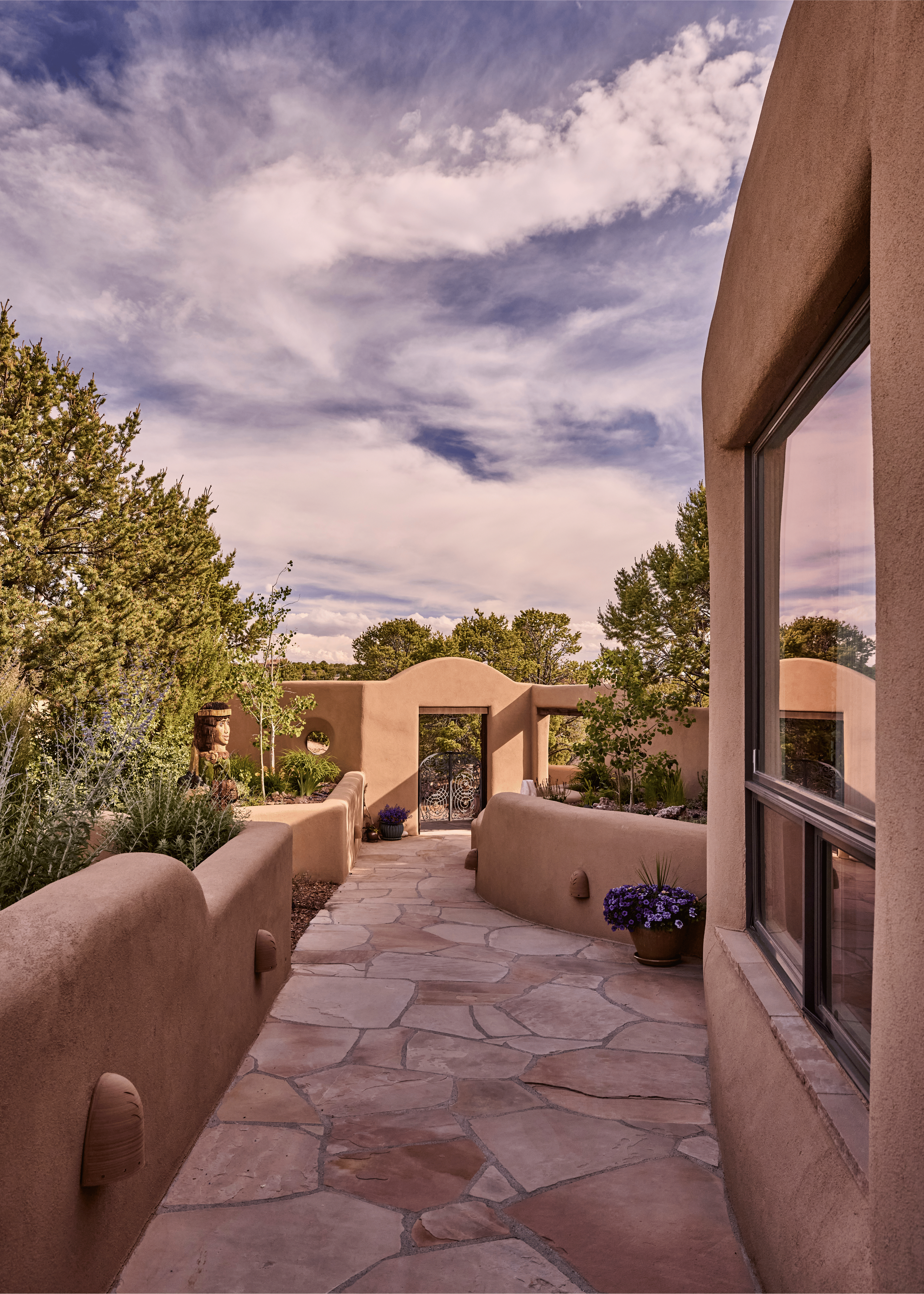 The approach to the Mesa Project: a flagstone path curves between adobe walls planted with native desert flora, leading to the ornate iron gate at the entry. A large carved sculptural figure stands sentinel among the plantings - the kind of detail th