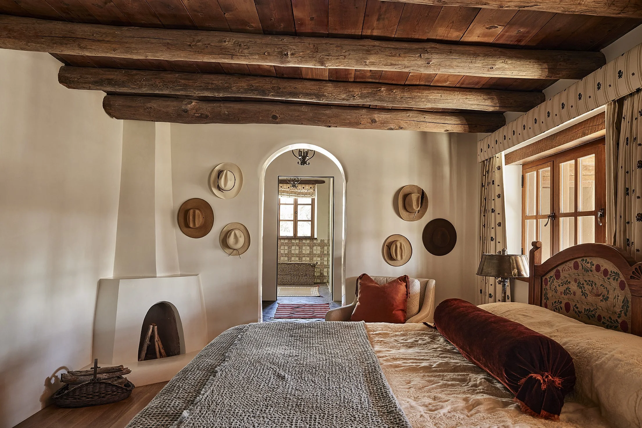 A warm Southwestern-style bedroom with a white kiva fireplace, exposed beams, and a wall decorated with cowboy hats, viewed toward an arched doorway.