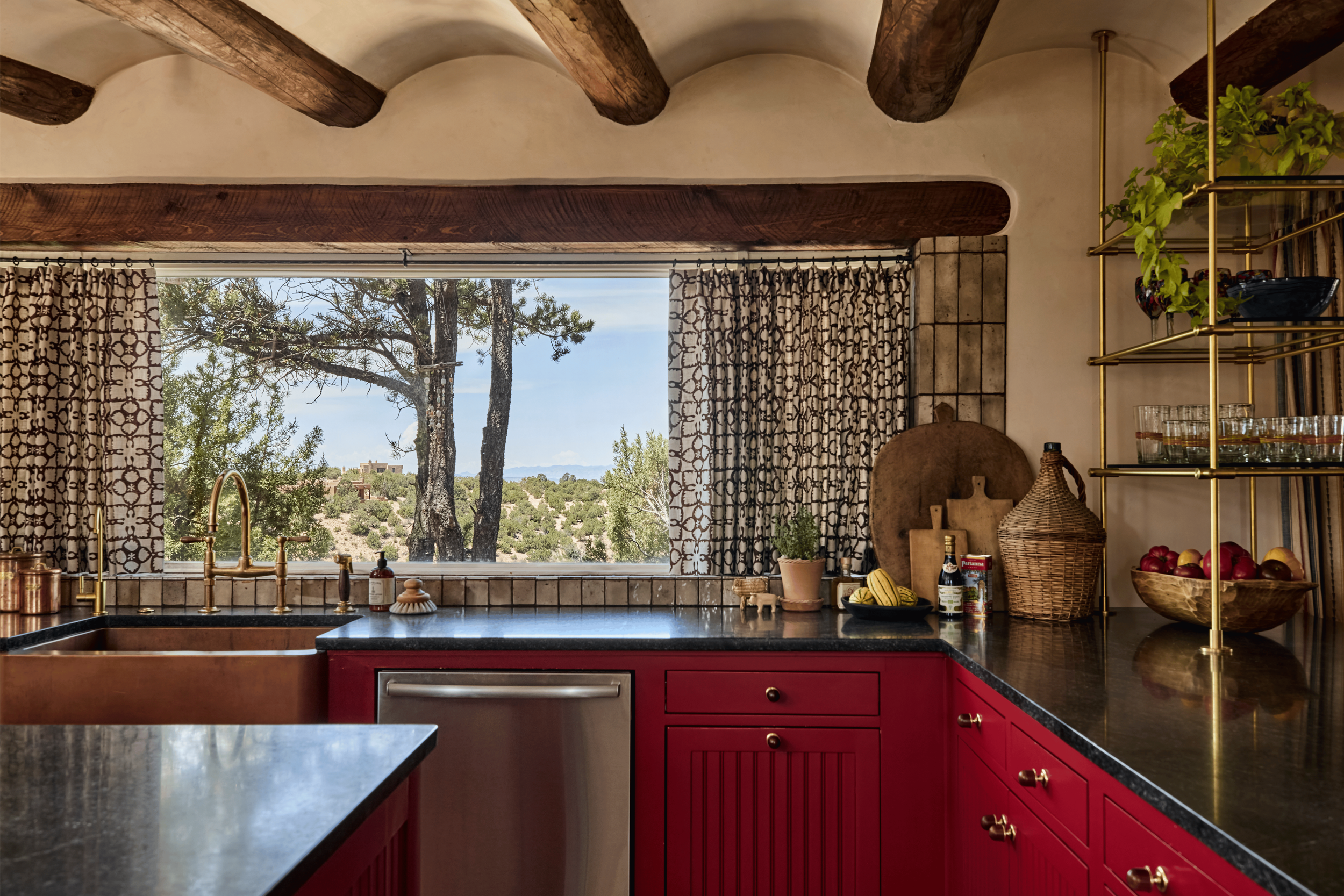 The kitchen sink wall at the Mesa Project frames the high desert through a picture window - pinon trees and mesa beyond a copper farmhouse sink, brass bridge faucet, and red beadboard cabinets. French & French Interiors designed this view into the ki