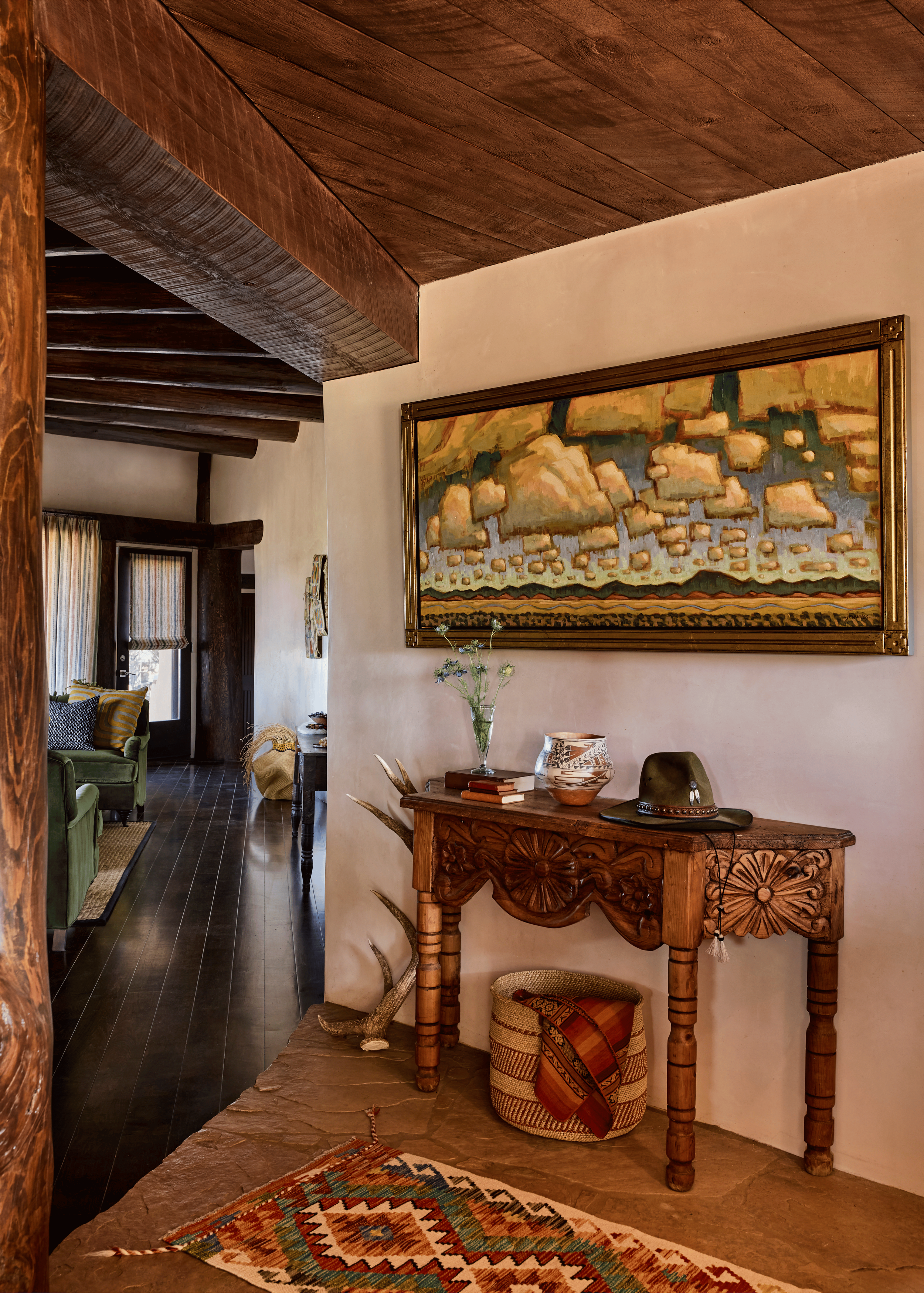 The entry hall of the Mesa Project announces the home's entire design language in a single frame: a carved wooden console, a large Southwest landscape painting, a rounded adobe arch leading to a terracotta-washed corridor beyond, and a kilim runner o