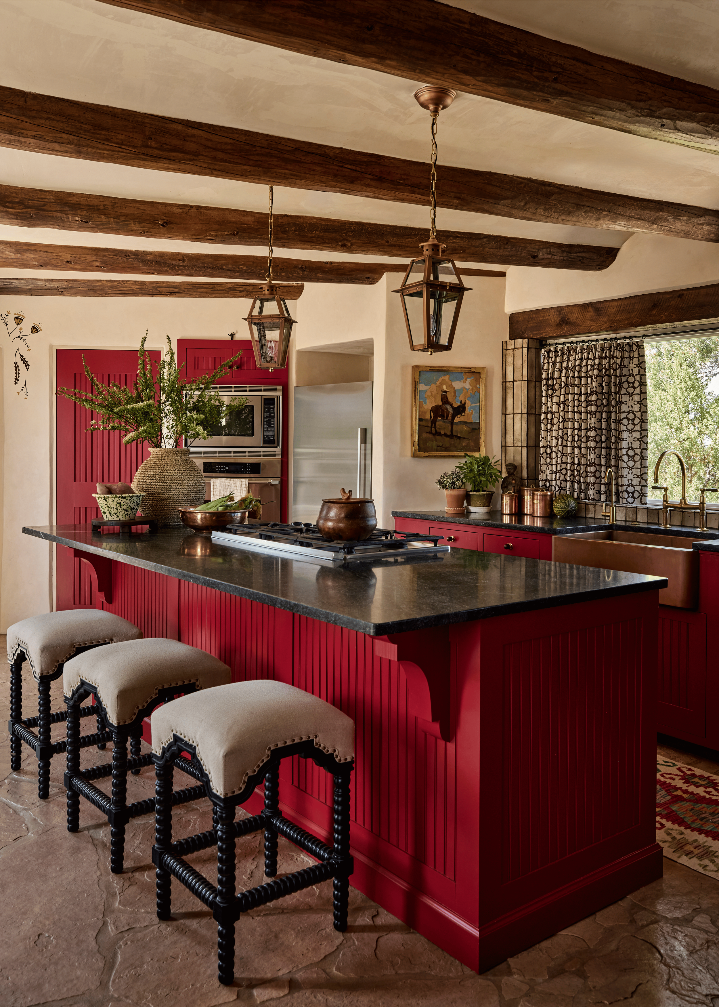 Aged brass lantern pendants hang over the kitchen island at the Mesa Project, where a gas cooktop sits alongside copper cookware and a small Southwest landscape painting anchors the wall behind. French & French Interiors designed this kitchen to be a