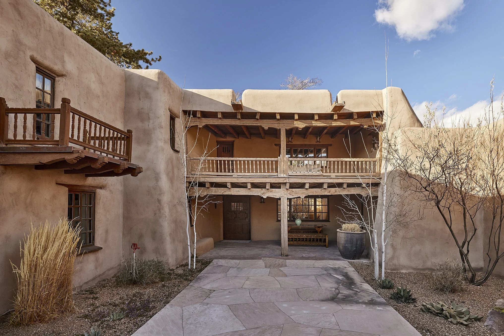 Traditional adobe-style courtyard with upper and lower balconies, wooden beams, and natural landscaping under a clear blue sky.