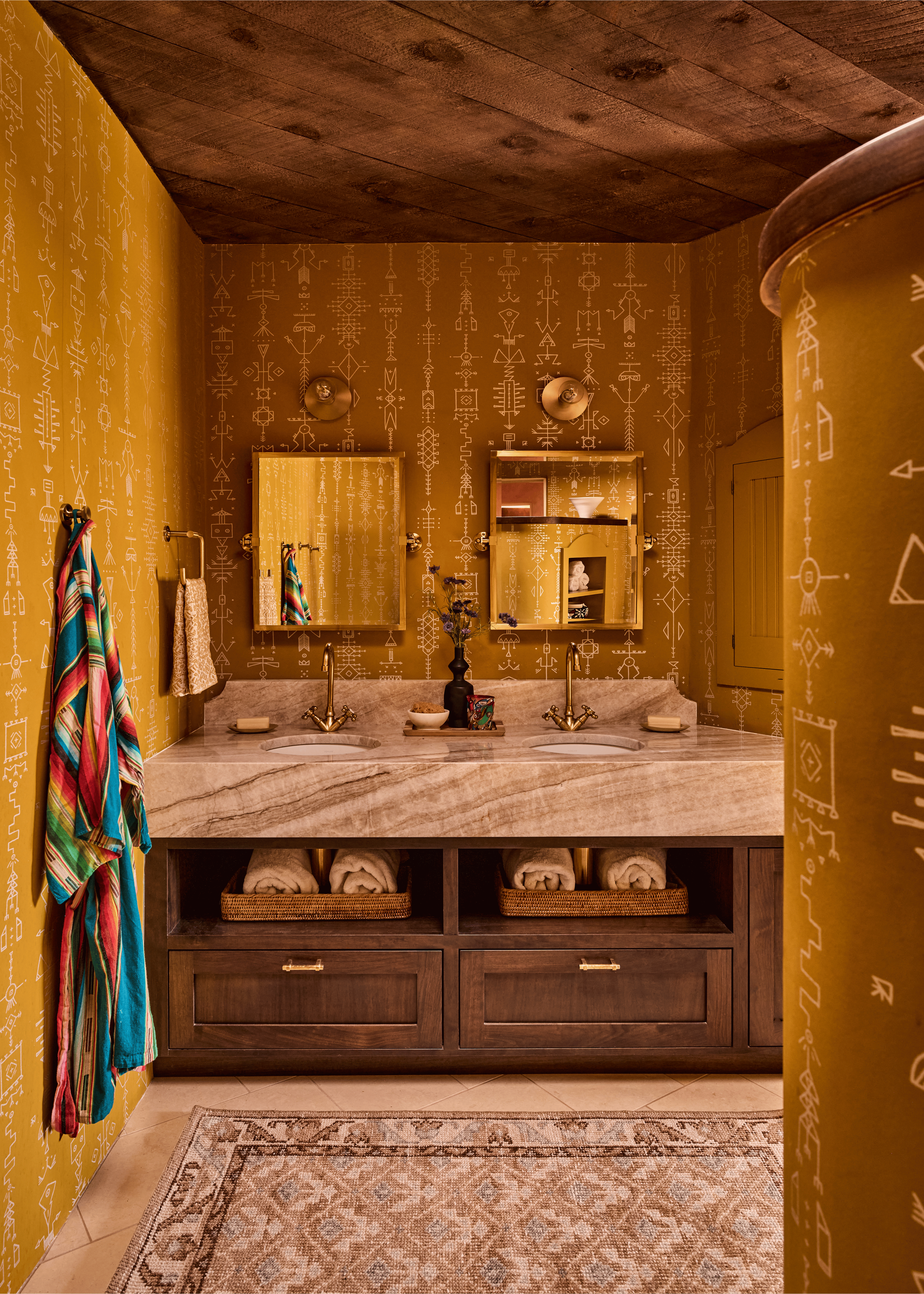 The primary bath at the Mesa Project makes no compromises: mustard tribal-print wallpaper wraps all four walls, a plank ceiling echoes the vigas throughout the home, and a marble double vanity with unlacquered brass anchors the space. Designed by Fre
