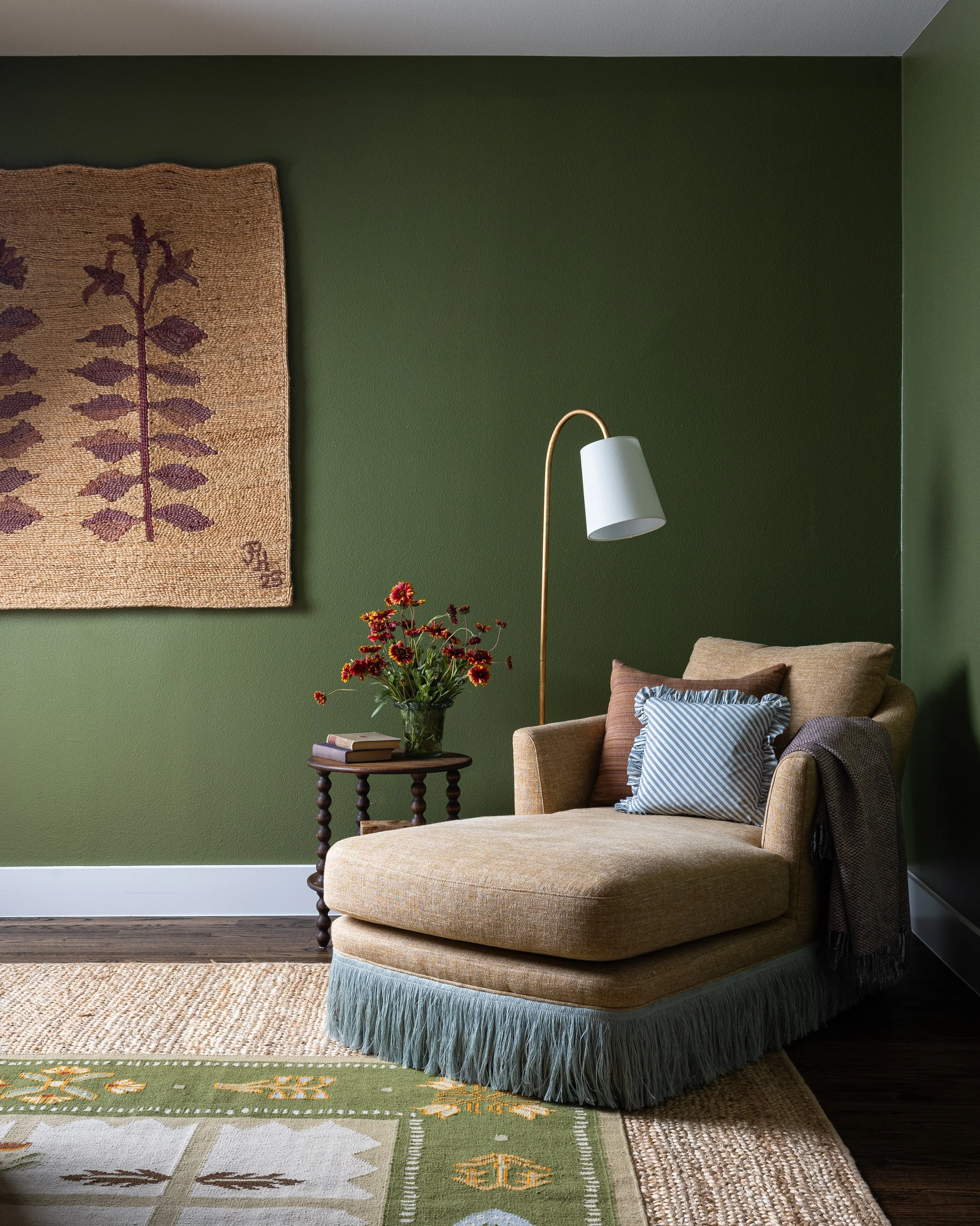 A reading nook in a Dallas cottage home - a fringed chaise, a vintage hooked rug hung as art, and gathered wildflowers on a bobbin table against deep forest green walls. Quiet luxury that feels genuinely personal.
