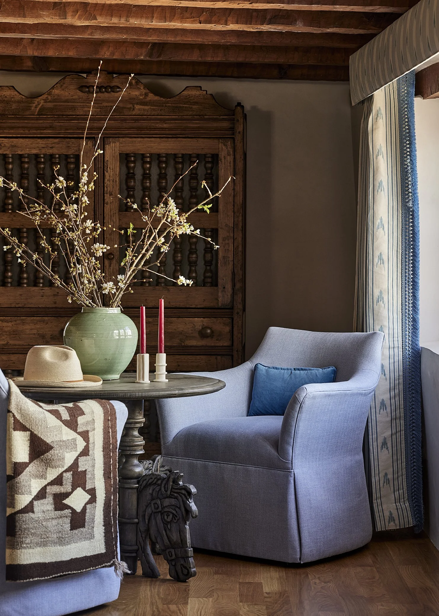 A small sitting nook featuring a soft blue armchair with a turquoise pillow, paired with a rustic round table holding a green vase with blossoming branches and red taper candles. A carved wooden cabinet with spindle details anchors the background.