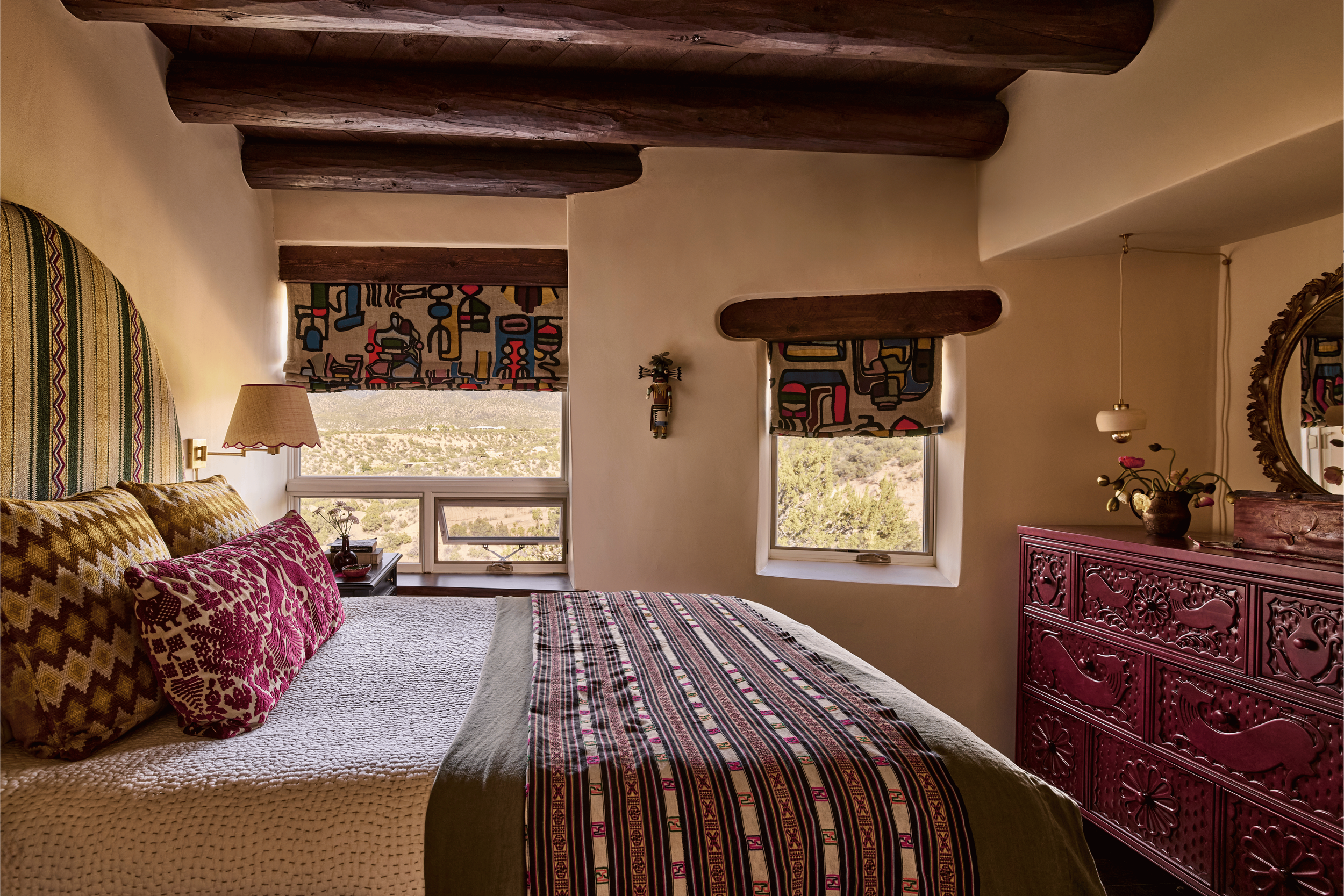 
Two windows frame the high desert in this guest bedroom at the Mesa Project - each dressed in bold abstract print roman shades and flanked by a small Kachina figure on the wall between them. The carved magenta dresser and green-striped headboard com