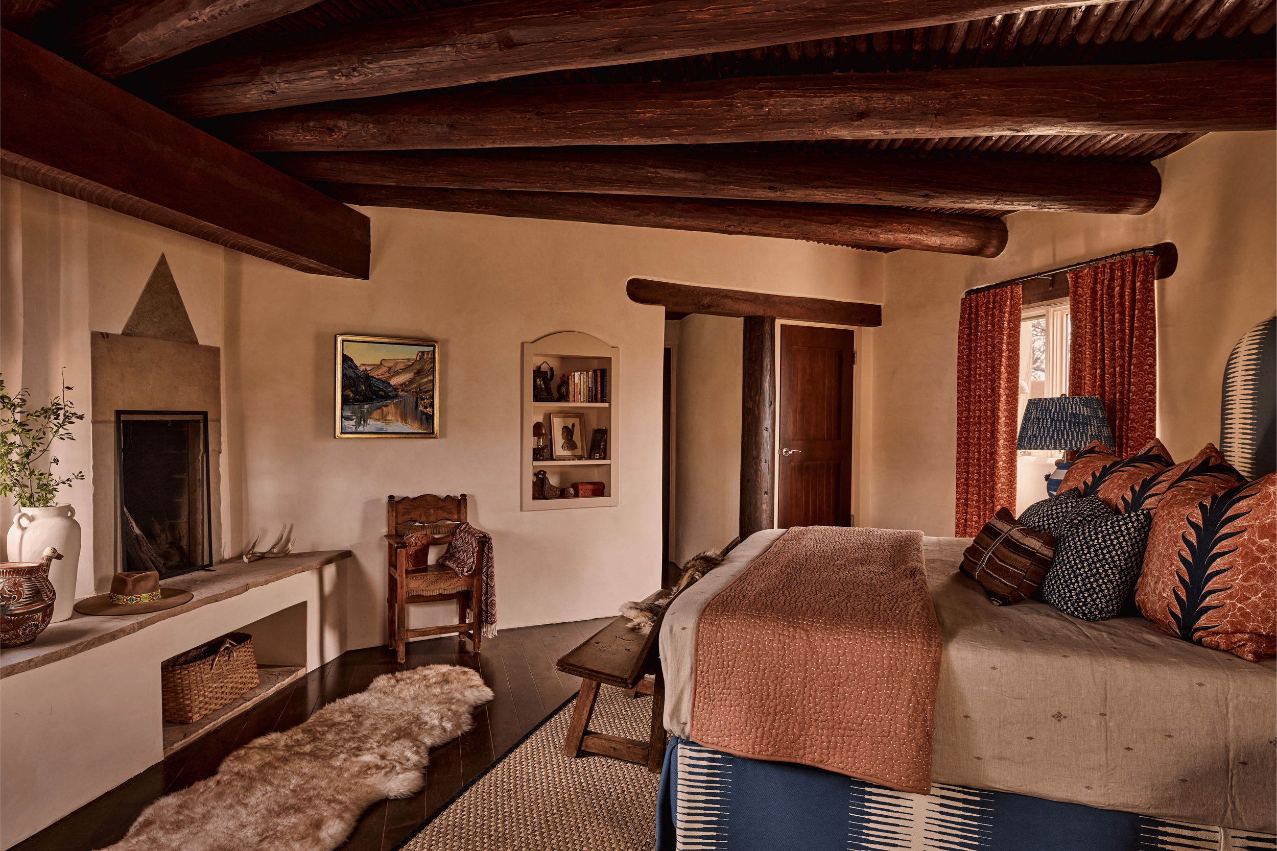 The primary bedroom at the Mesa Project has the quiet authority of a room that has been lived in well: a kiva fireplace with a carved stone surround, built-in arched bookshelves, an antique carved chair, and rust block-print drapes - all designed by 