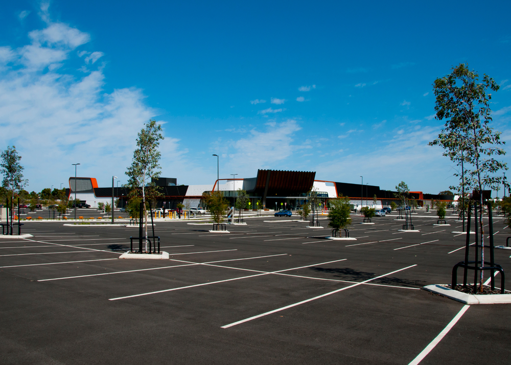 Empty parking lot with newly planted trees under a bright blue sky, in front of a low building.