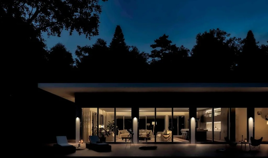 Exterior night view of a luxury modern residence in Girona, featuring a flat roof design, floor-to-ceiling glass walls illuminated from within, and a dark forest backdrop, designed by Sebastian PM.