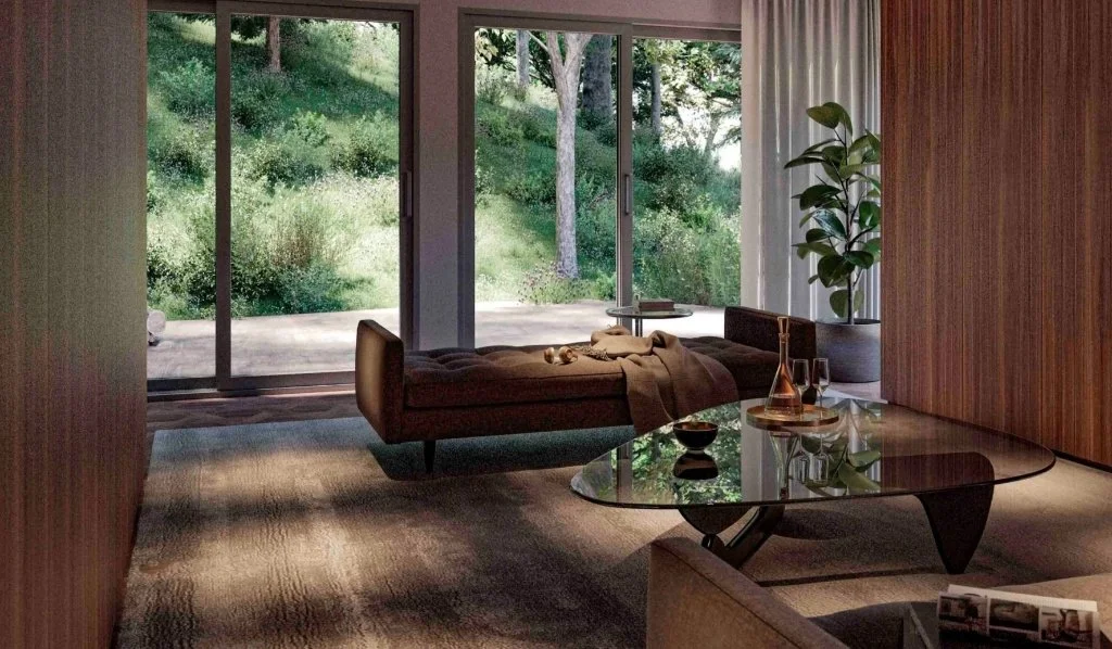 Modern biophilic lounge area in a Girona forest house, featuring a brown tufted daybed, a smoked glass coffee table, and large sliding glass doors overlooking the lush woodland, designed by Sebastian PM.