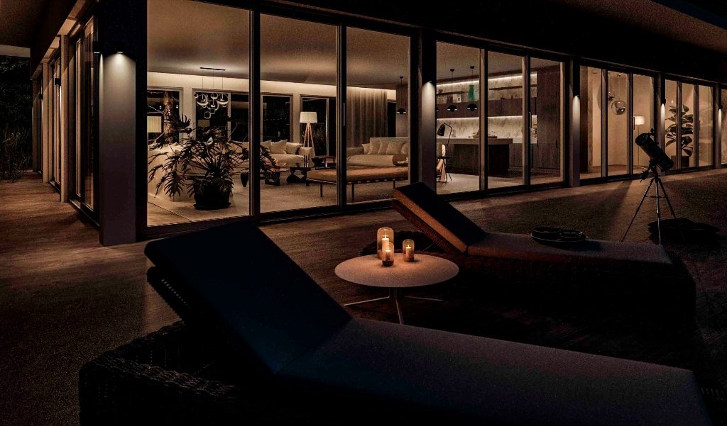 Outdoor terrace of a luxury Girona residence at night, featuring designer sun loungers, candle lighting, and a panoramic view of the illuminated open-plan interior through glass walls, by Sebastian PM.