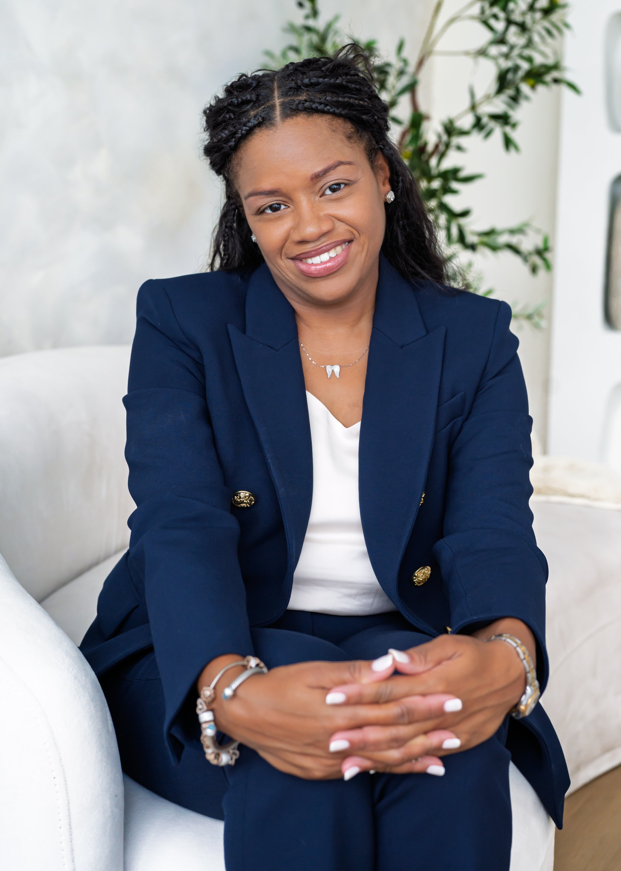 Sowania Germain, LMHC, smiling in a navy suit with her hands clasped, seated on a white chair