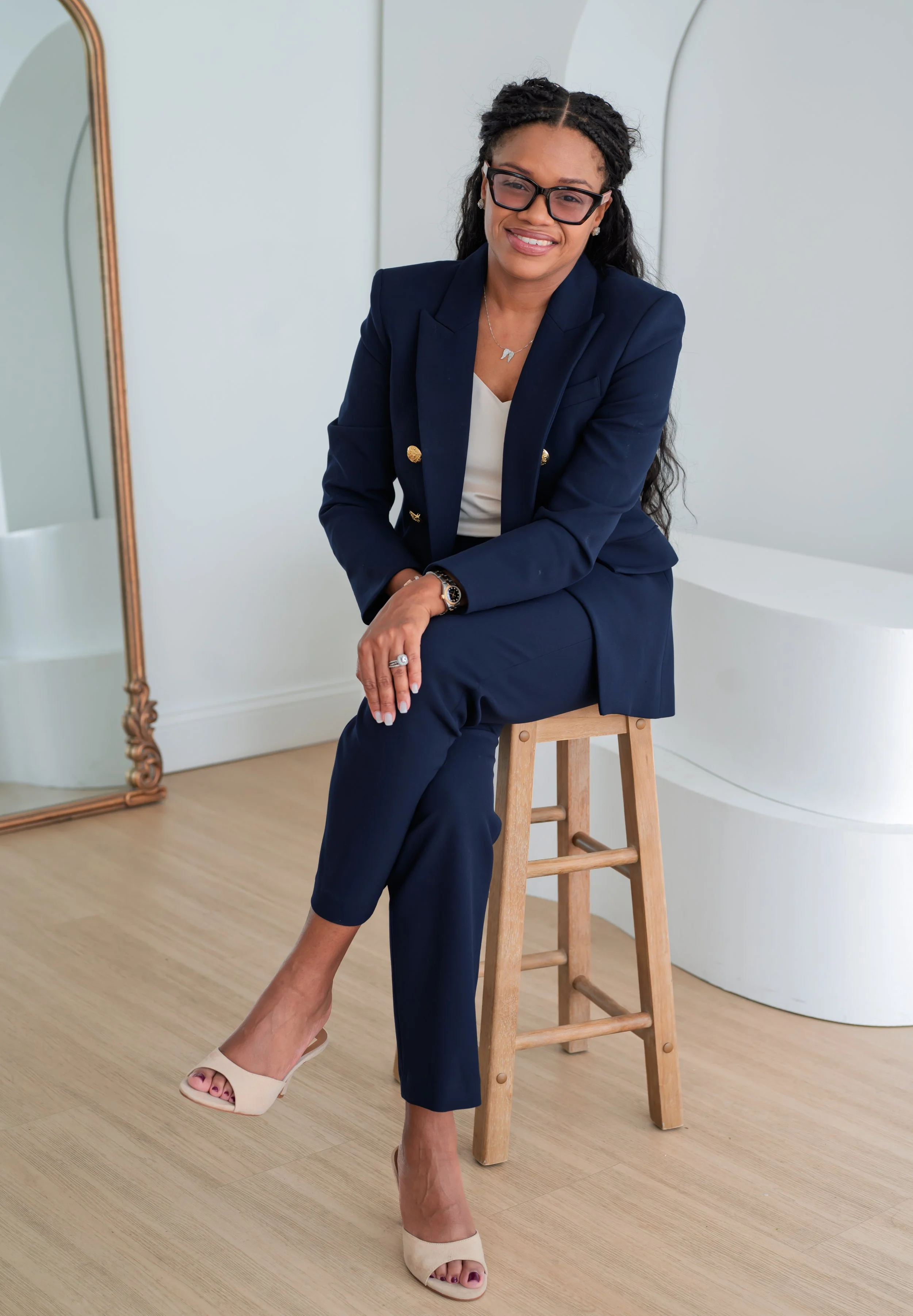 Sowania Germain, perinatal therapist in West Palm Beach, seated on a wooden stool in a navy suit and glasses
