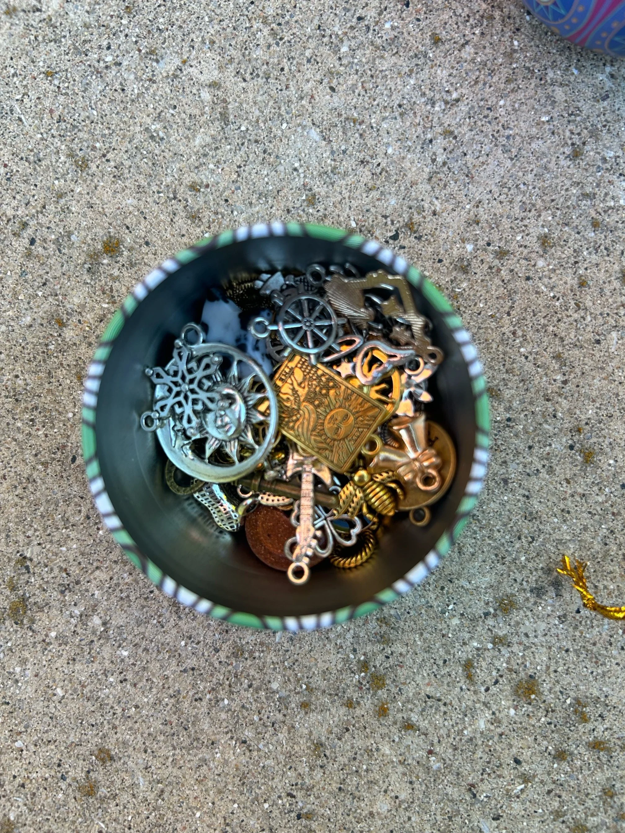 Top-down view of a jar filled with assorted small jewelry pieces, including gear charms, snowflake designs, gold-colored charms, and other metal trinkets, placed on sandy surface.