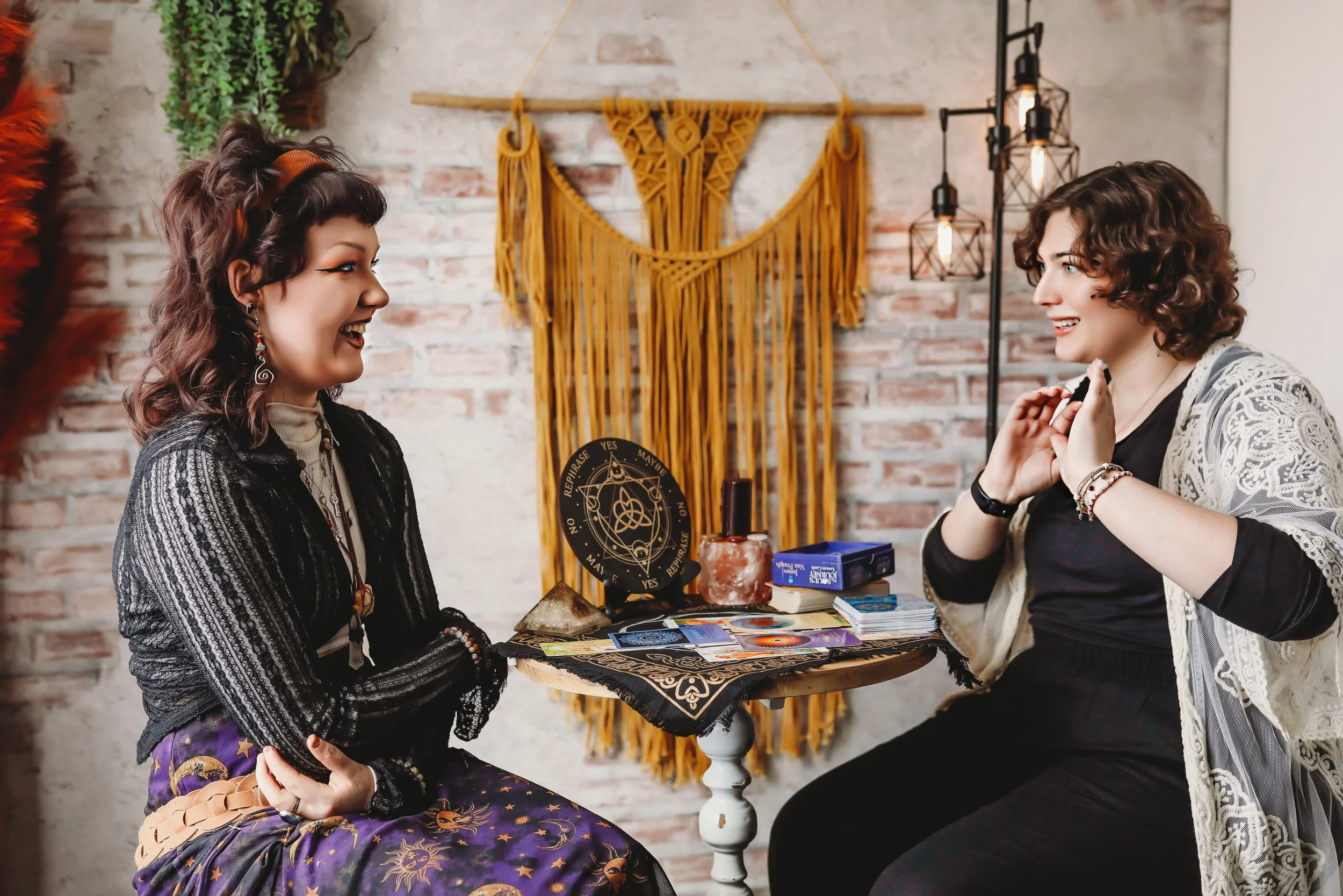 Two women sitting at a table in a cozy room with a brick wall, engaging in a tarot card reading, with mystical items and a yellow macrame wall hanging in the background.