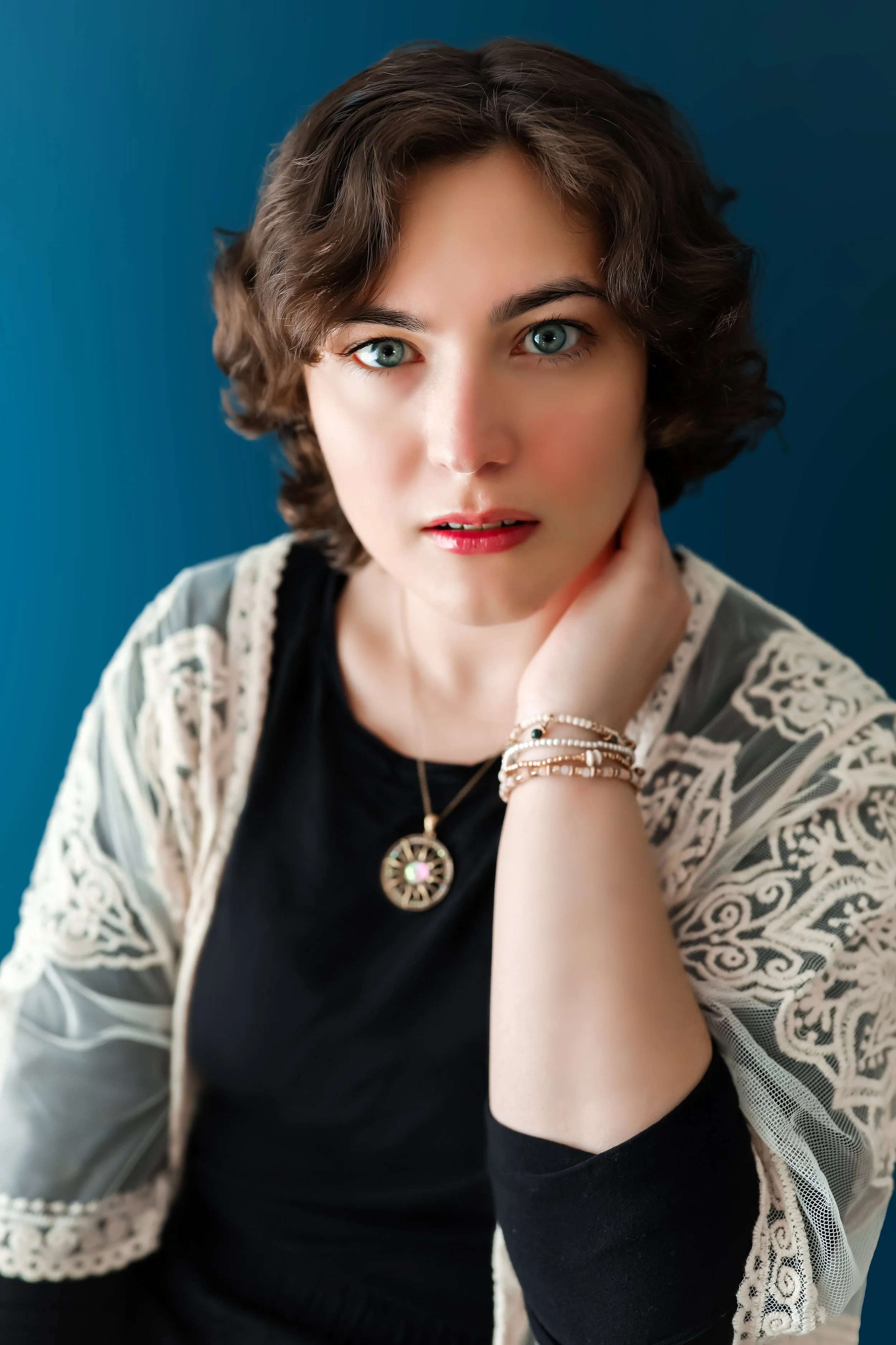 A woman with short curly brown hair and blue eyes looking directly at the camera, resting her chin on her hand. She is wearing a black top, layered jewelry including a round pendant necklace, and bracelets, with a cream-colored patterned lace jacket against a blue background.
