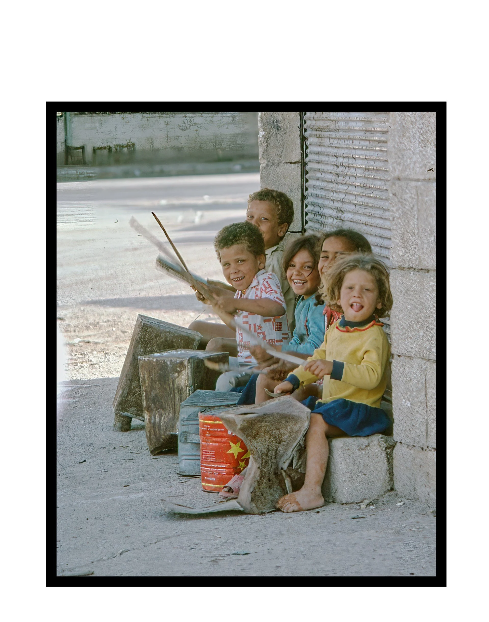 five children smiling and drumming on buckets and logs on a street corner