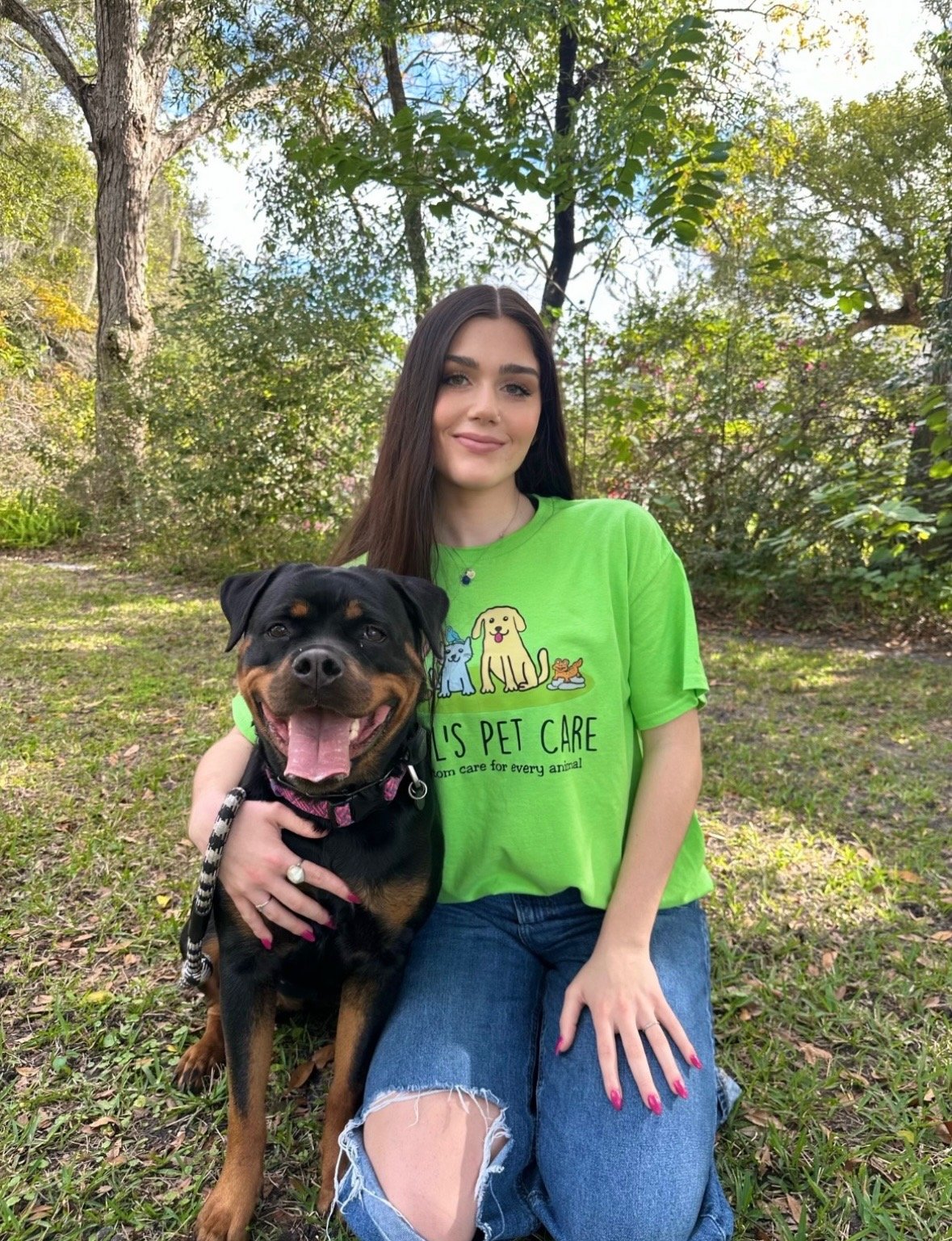 A young woman with long dark hair, wearing a bright green t-shirt featuring cartoon animals and the text 'LIS PET CARE', sitting on grass with a black and tan dog that has a pink collar, outdoors among trees and bushes on a sunny day.