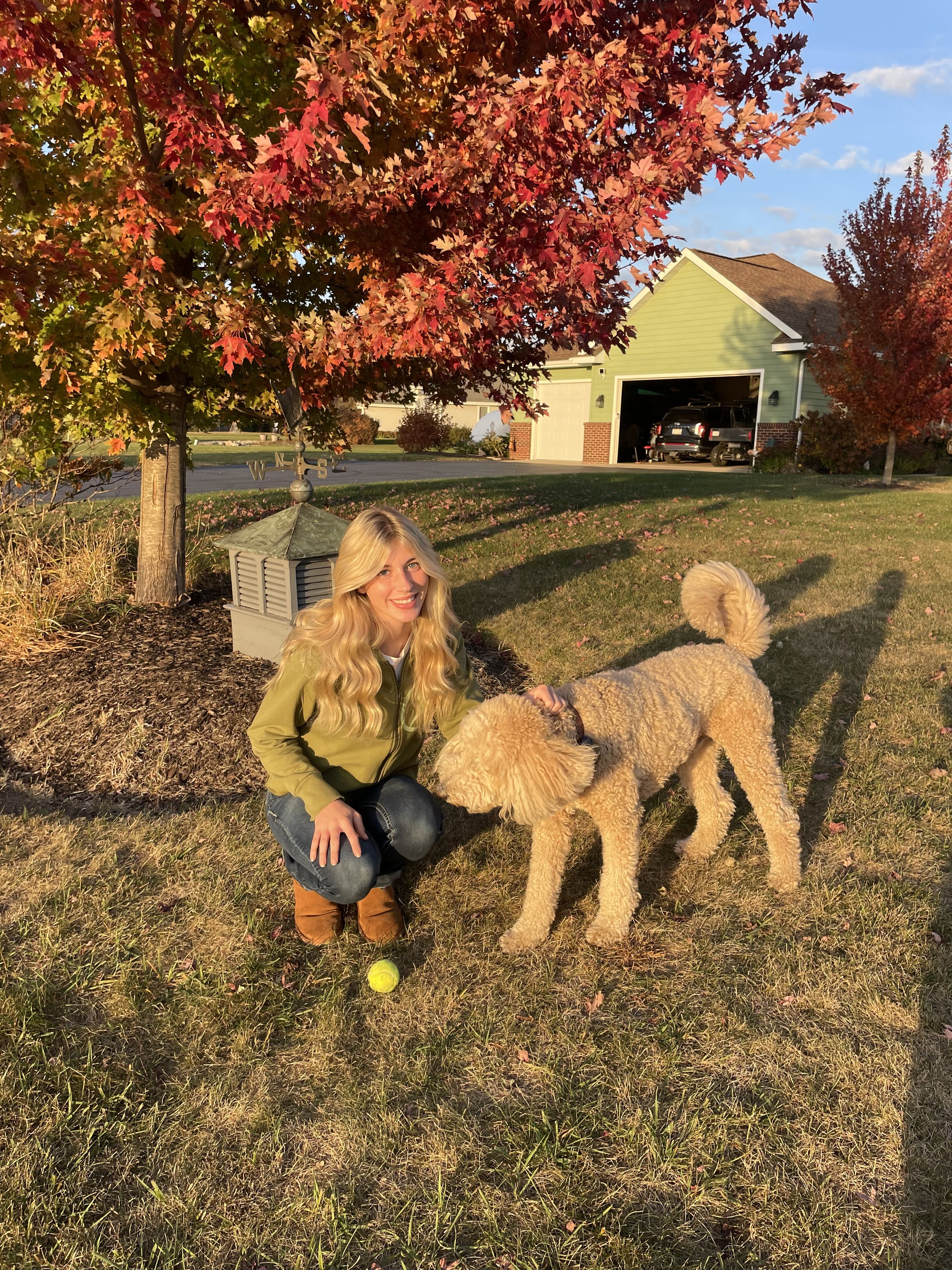A woman with long blonde hair, smiling, kneeling on the grass next to a light-colored poodle. They are outdoors on a sunny day, near a tree with red leaves, a house with a garage, and a car in the background. There's a tennis ball on the grass near the woman.
