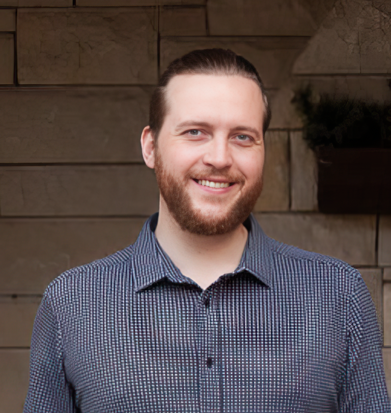 A smiling man with a beard and slicked-back hair in a blue patterned shirt, standing outdoors in front of a wooden wall.