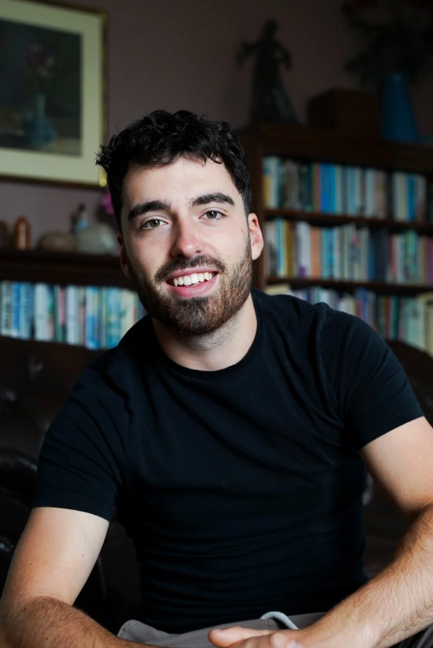 A young man with dark hair and a beard, smiling, wearing a black t-shirt, sitting in a room with a bookshelf filled with books behind him.