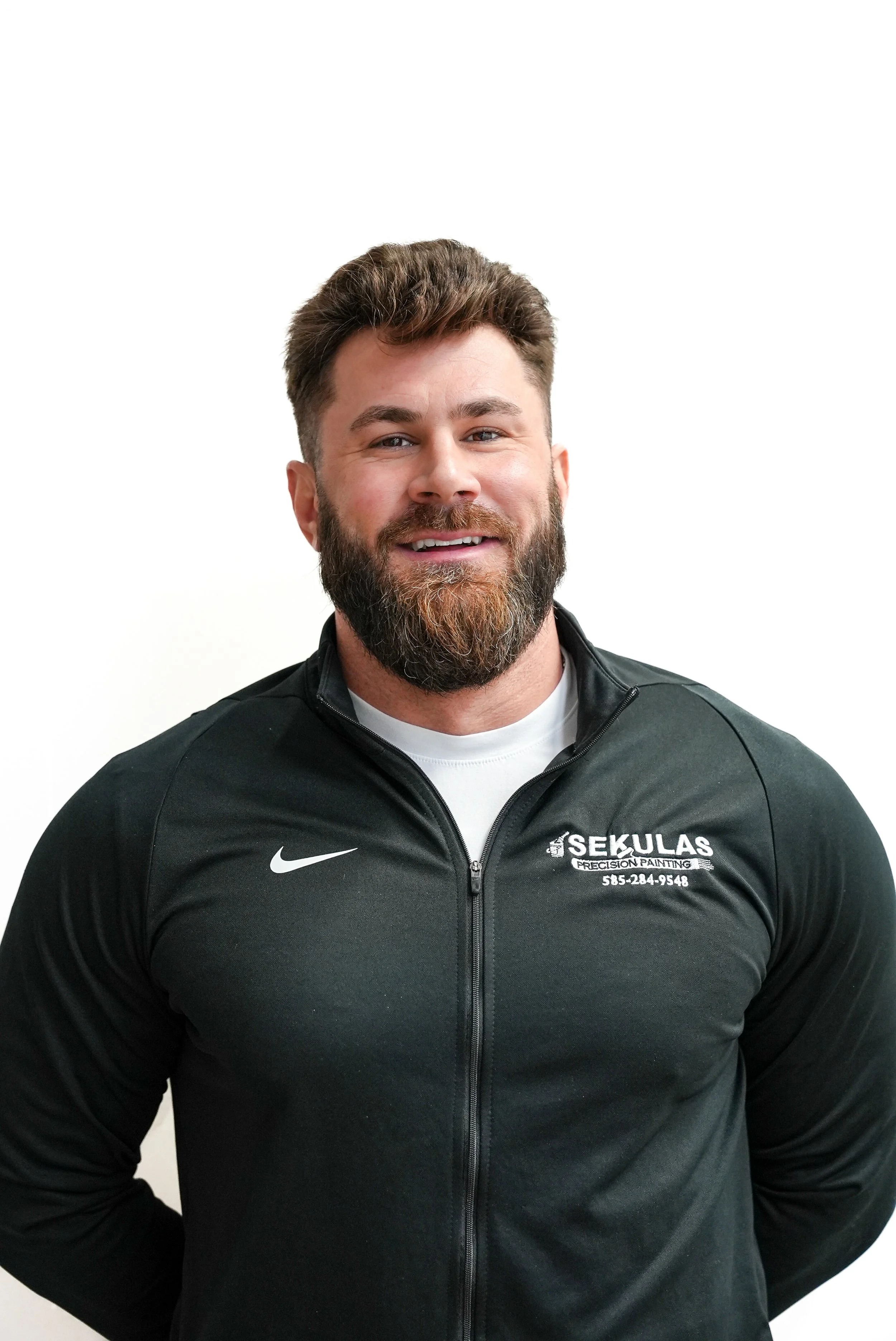 A smiling man with a beard and brown hair wearing a black jacket with a logo that says 'SEKULAS PRECISION PAINTING' and a phone number, standing against a white background.