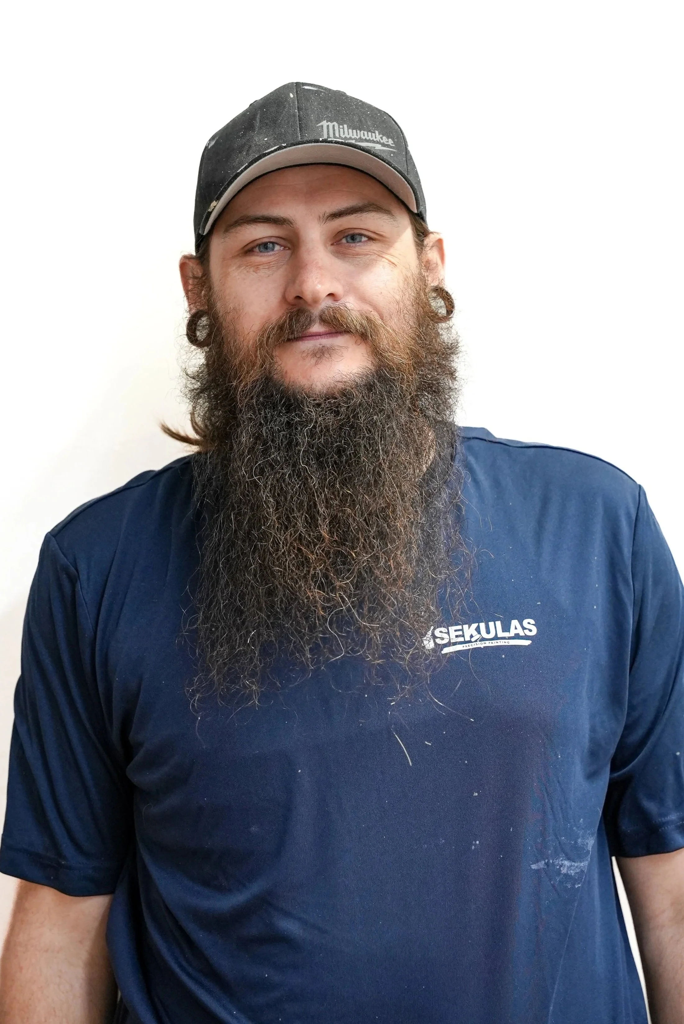 A man with a long beard, mustache, and stretched earlobes wearing a dark cap with 'Milwaukee' written on it and a blue T-shirt with the word 'SEKULAS' on it, standing against a plain white background.