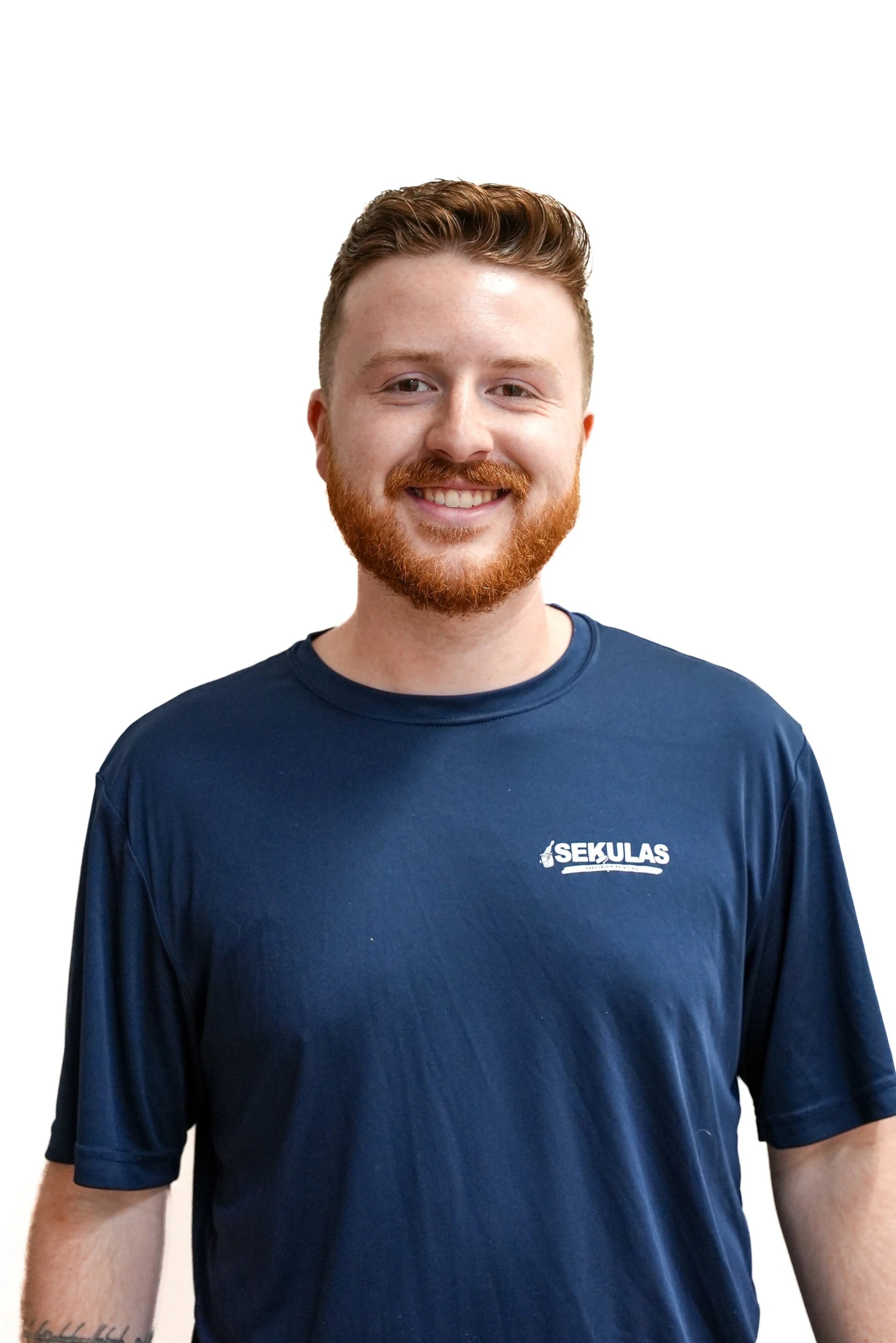 A young man with red hair and a beard smiling, wearing a navy blue shirt with the white 'SEKULAS' logo.