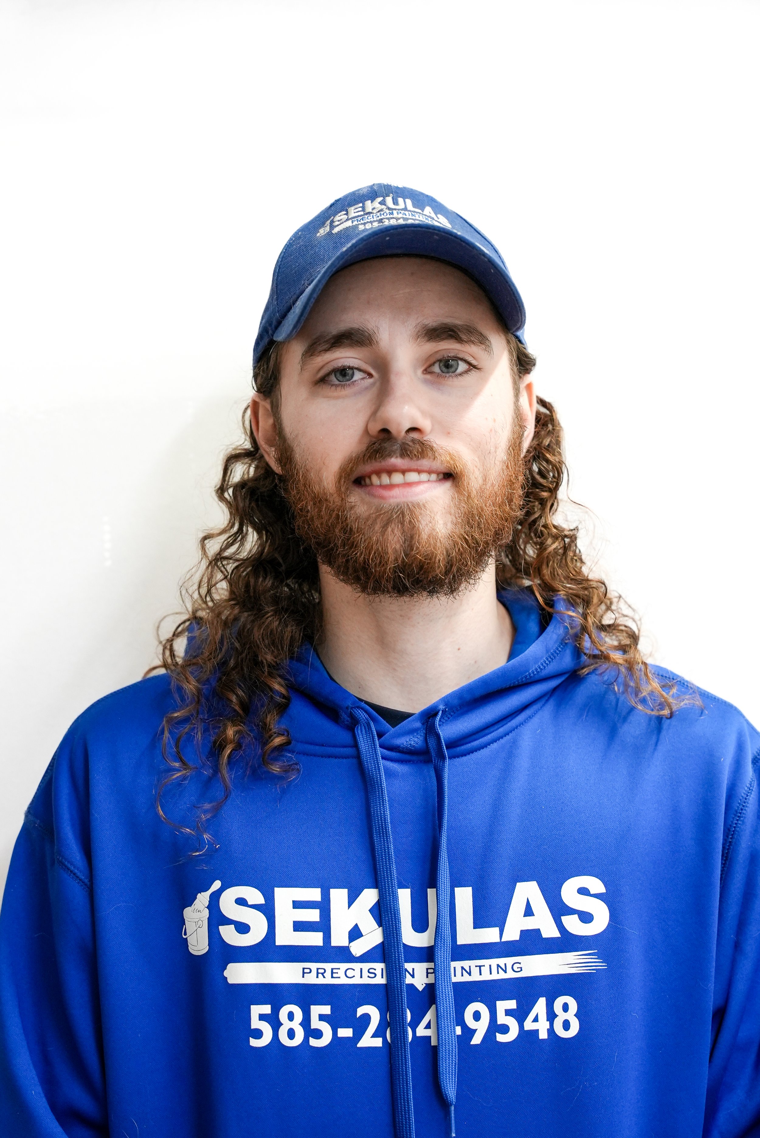 Young man with long curly hair and beard wearing a blue SEKULAS hoodie and baseball cap, standing against a white wall.