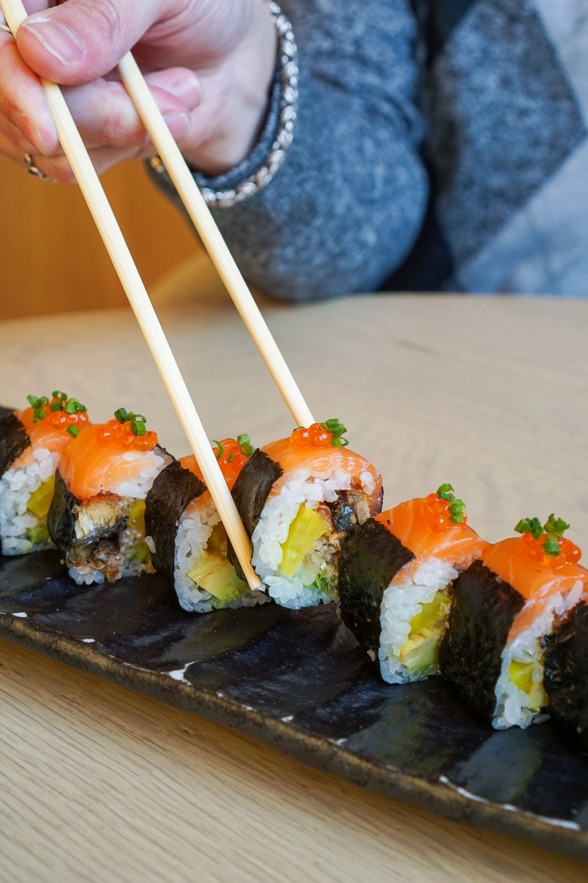A person using chopsticks to pick up sushi rolls on a black platter, with visible salmon, avocado, rice, and seaweed.