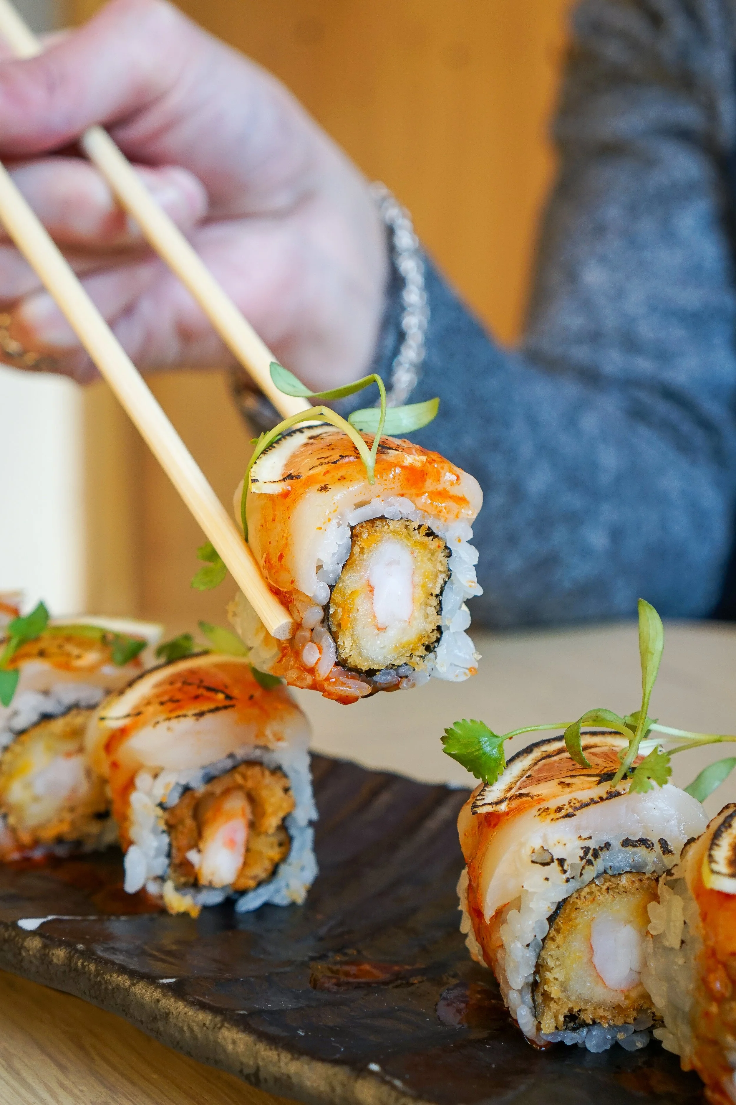 Close-up of a person holding chopsticks and picking up a sushi roll with a seafood filling, garnished with microgreens, on a black platter. Other sushi rolls are visible on the platter.
