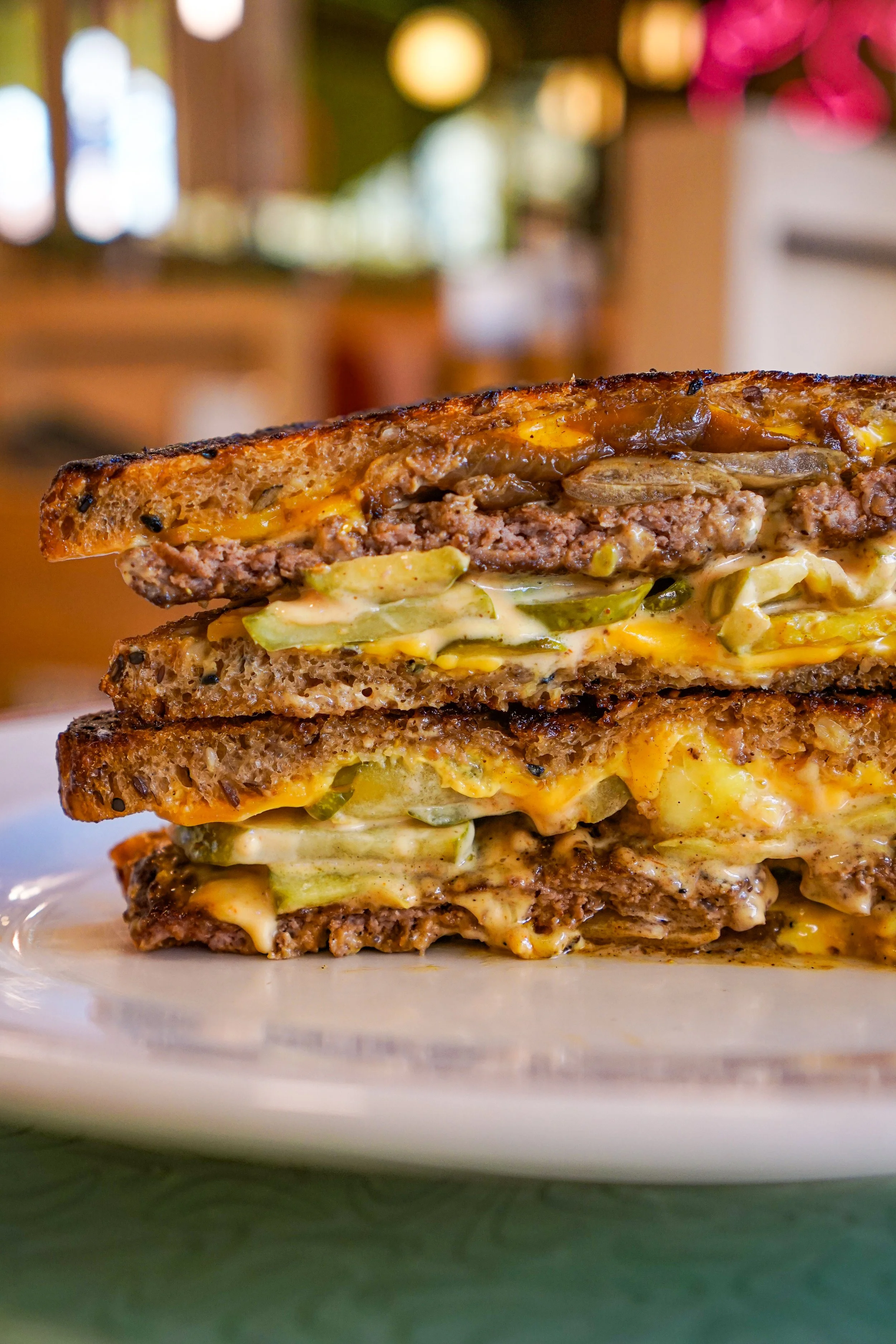 Close-up of a triple layered cheeseburger with bacon, lettuce, and melted cheese on toasted bread, served on a white plate. The background is blurred with warm lighting.