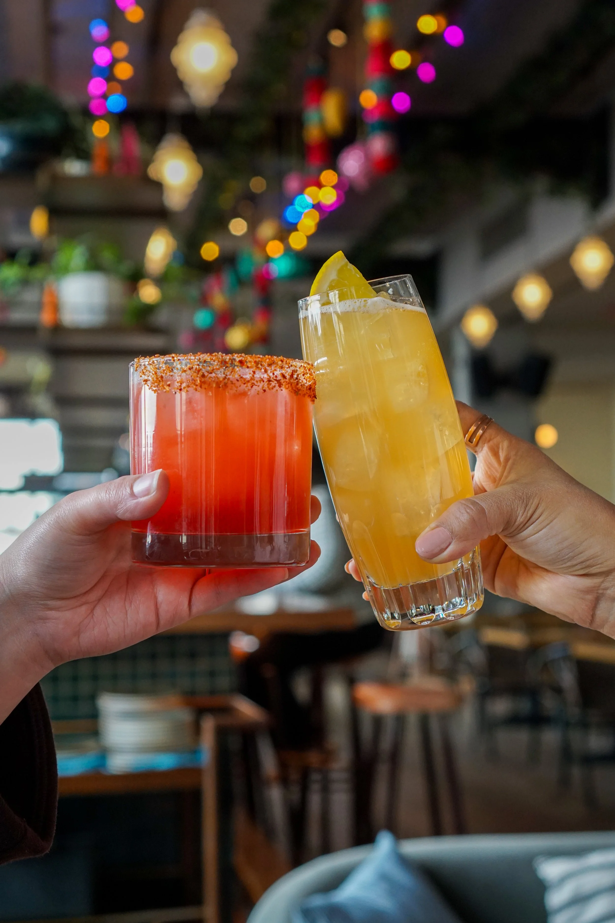 Two people holding up colorful cocktails in a festive setting with blurred lights and decorations in the background.