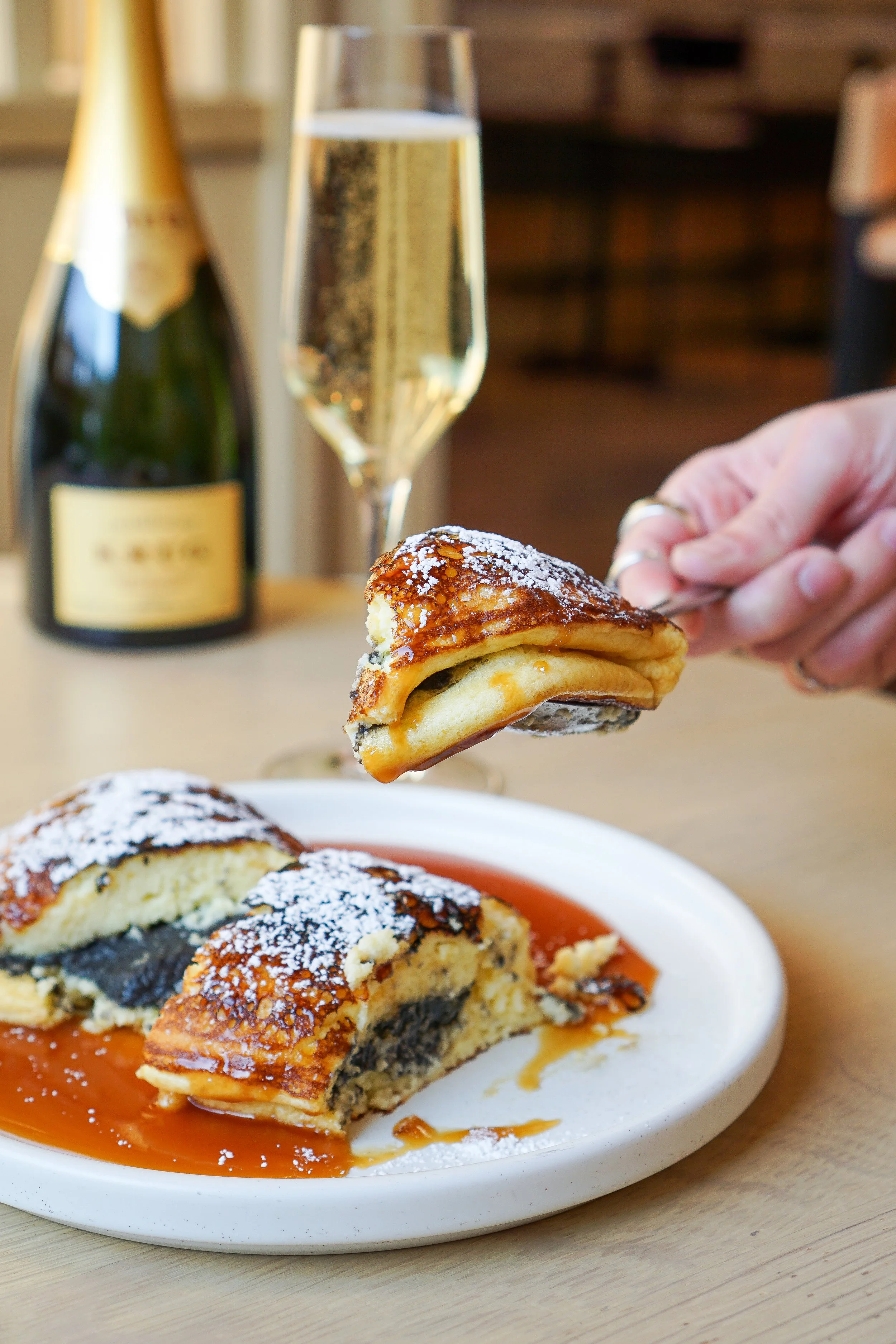 A plate with half-eaten poppy seed roll, a hand holding a piece of the pastry, a glass of champagne, and a champagne bottle in the background.