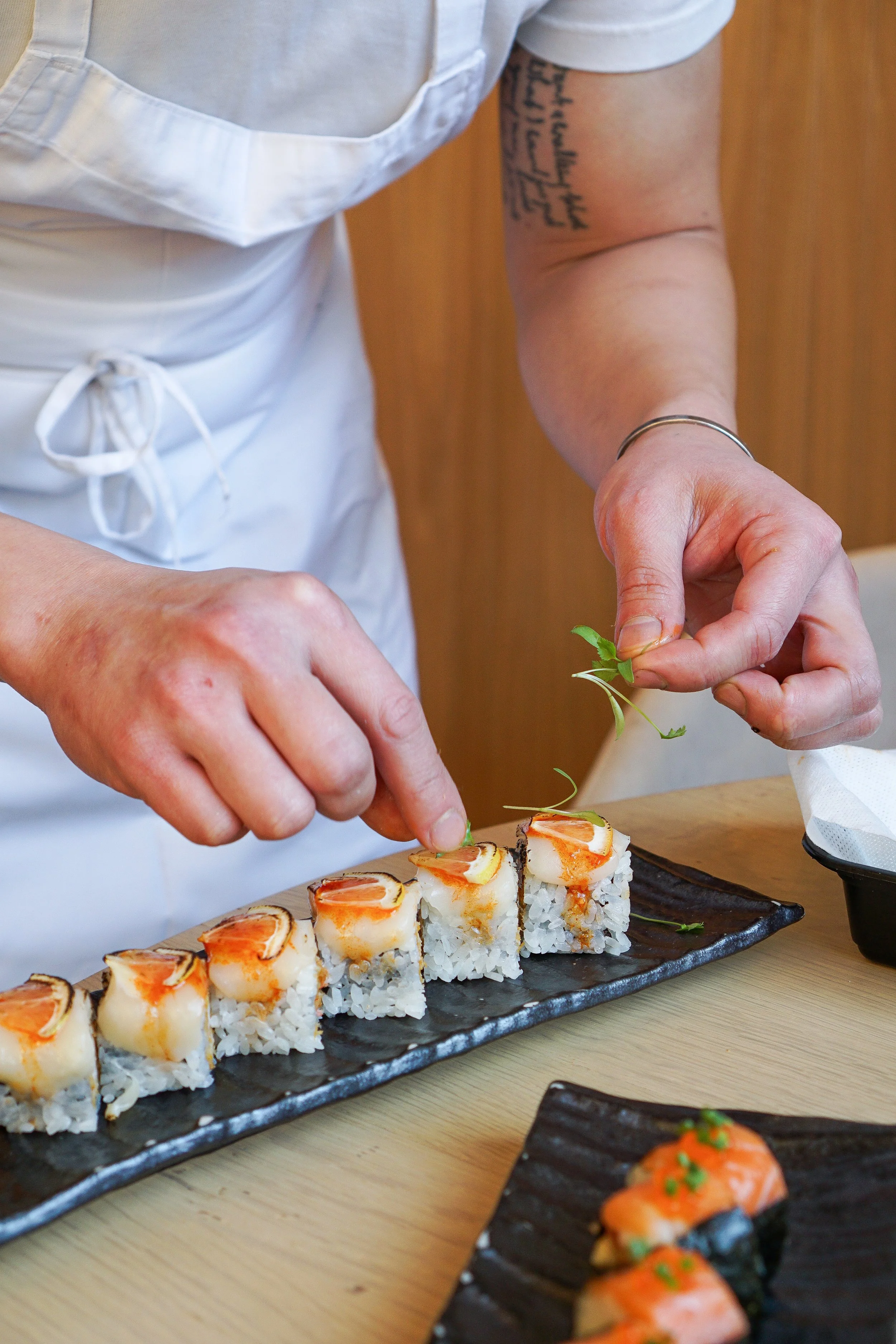 A chef garnishing sushi rolls with green herbs on a black rectangular plate.